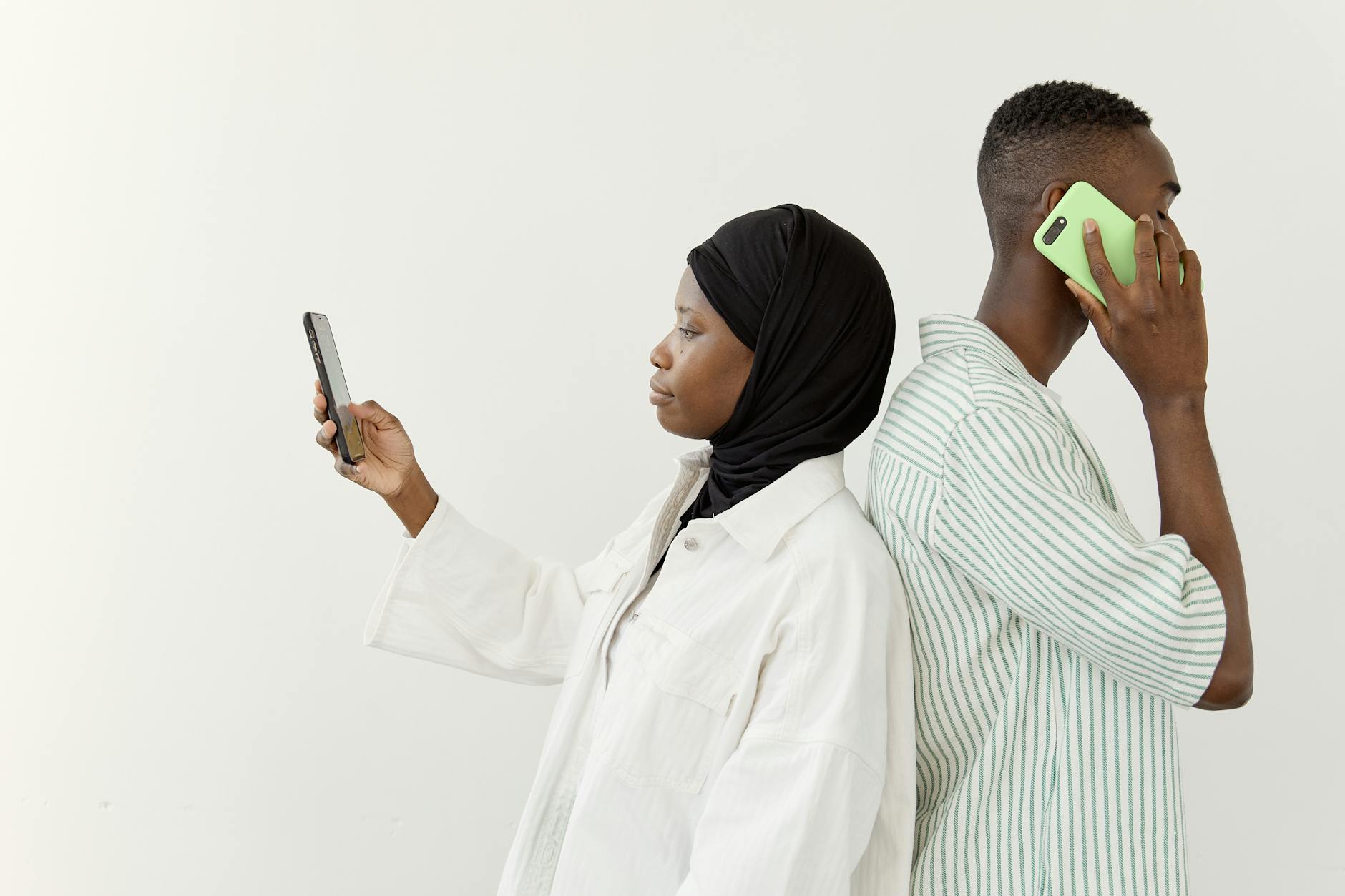 Muslim couple stands back-to-back, engaged with phones, illustrating modern communication.