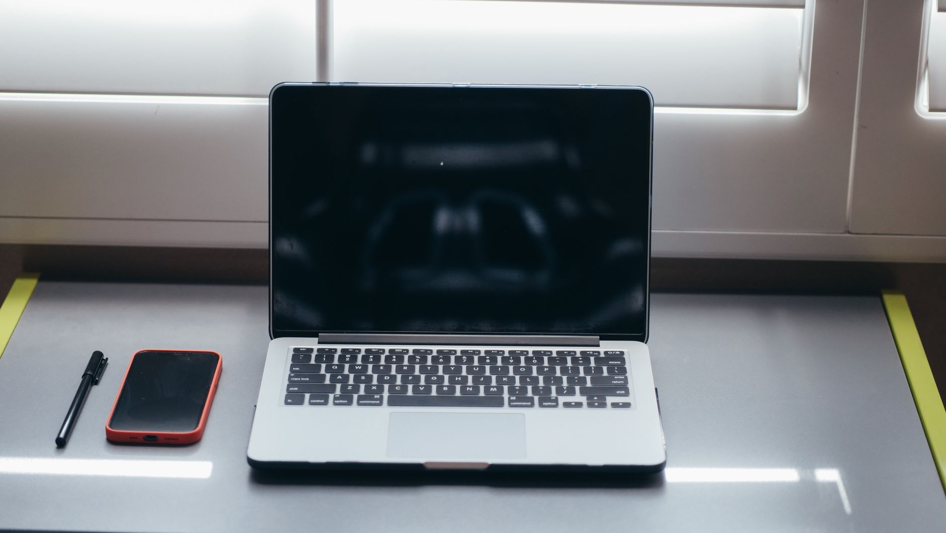 A minimalist desk setup featuring a laptop, smartphone, and pen under soft window light.