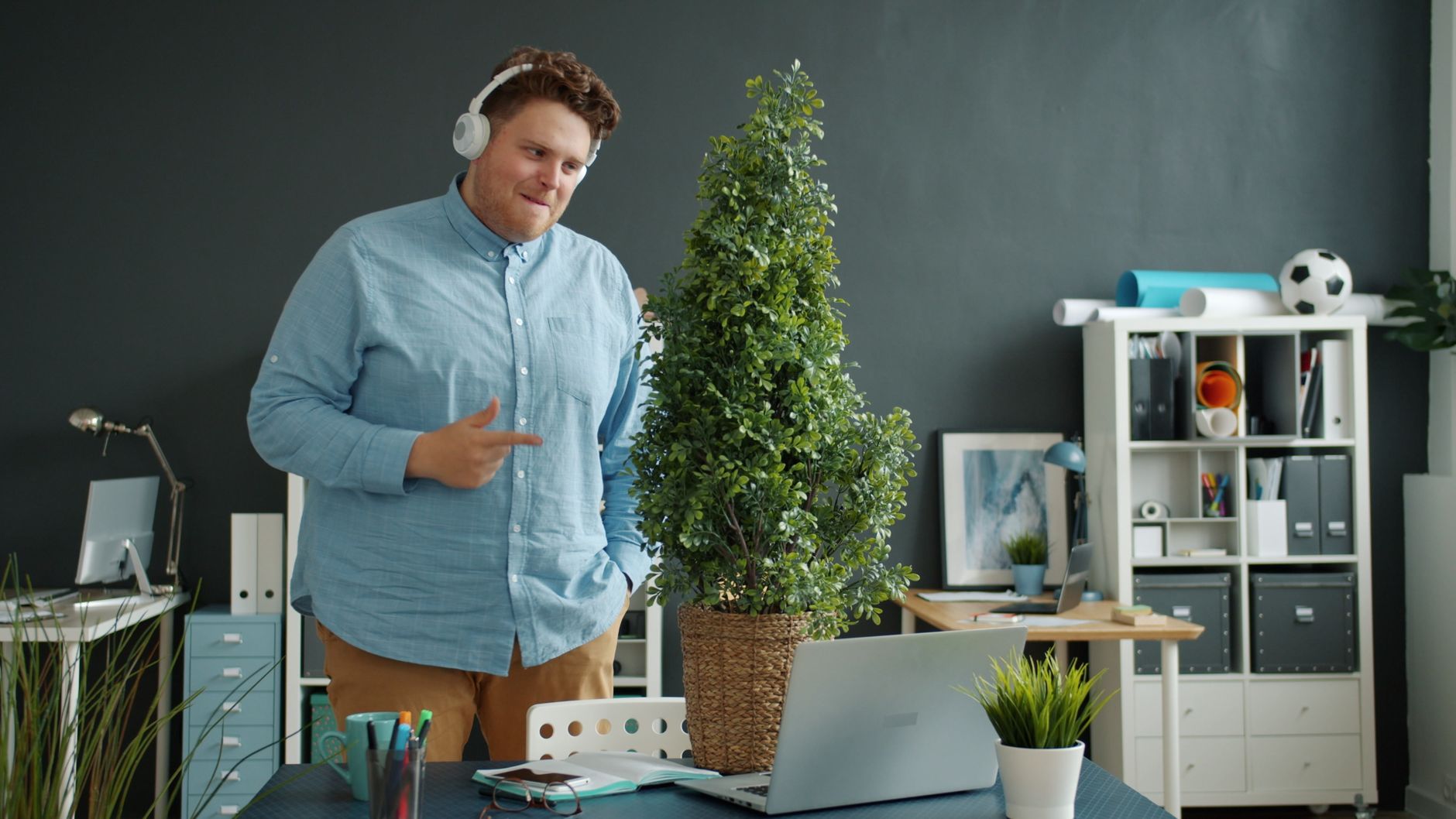 Man in modern office setting wearing headphones, standing by a large indoor plant.