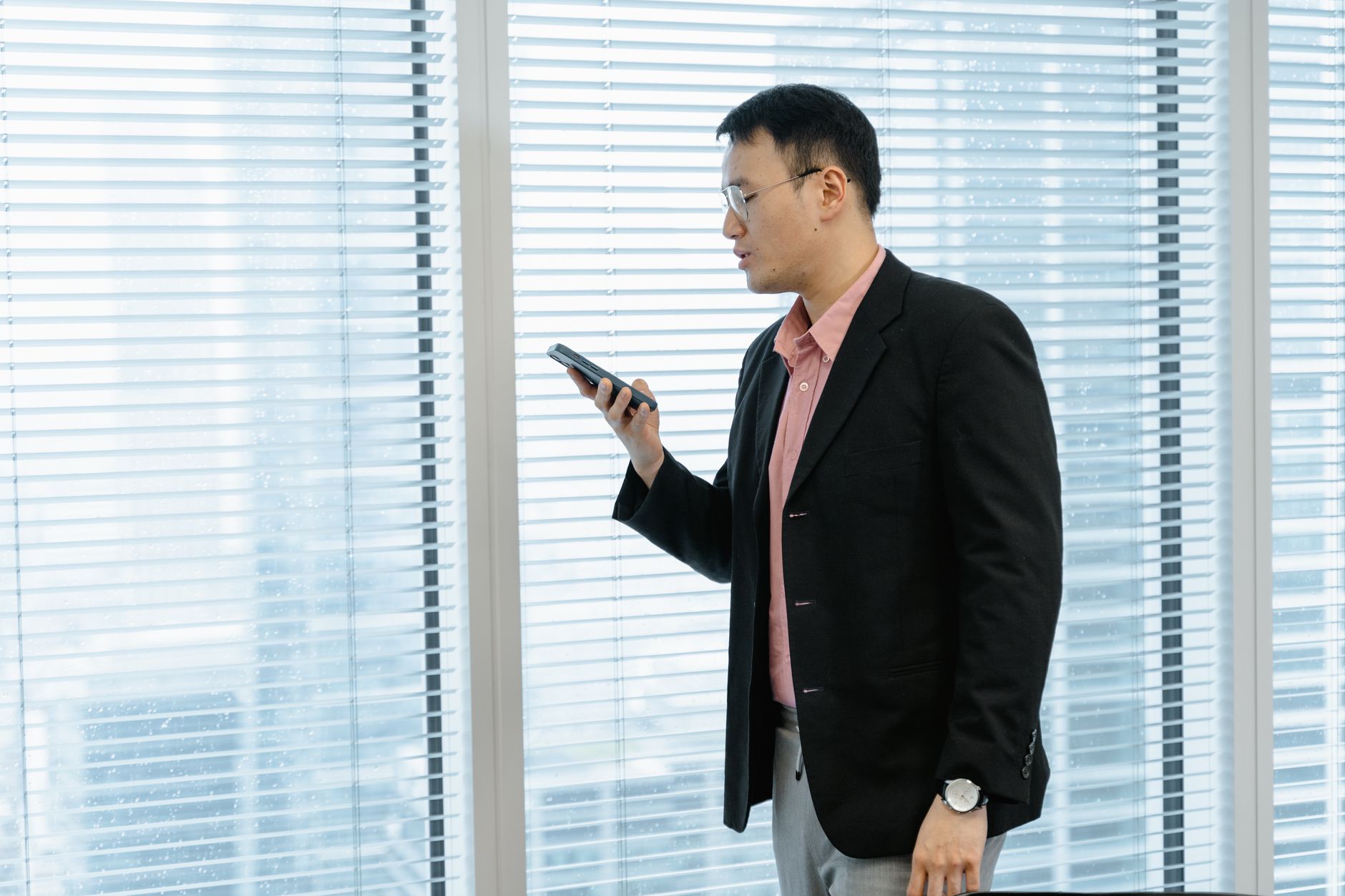 Asian man in office holding a mobile phone, appears to interact with the screen near window blinds.