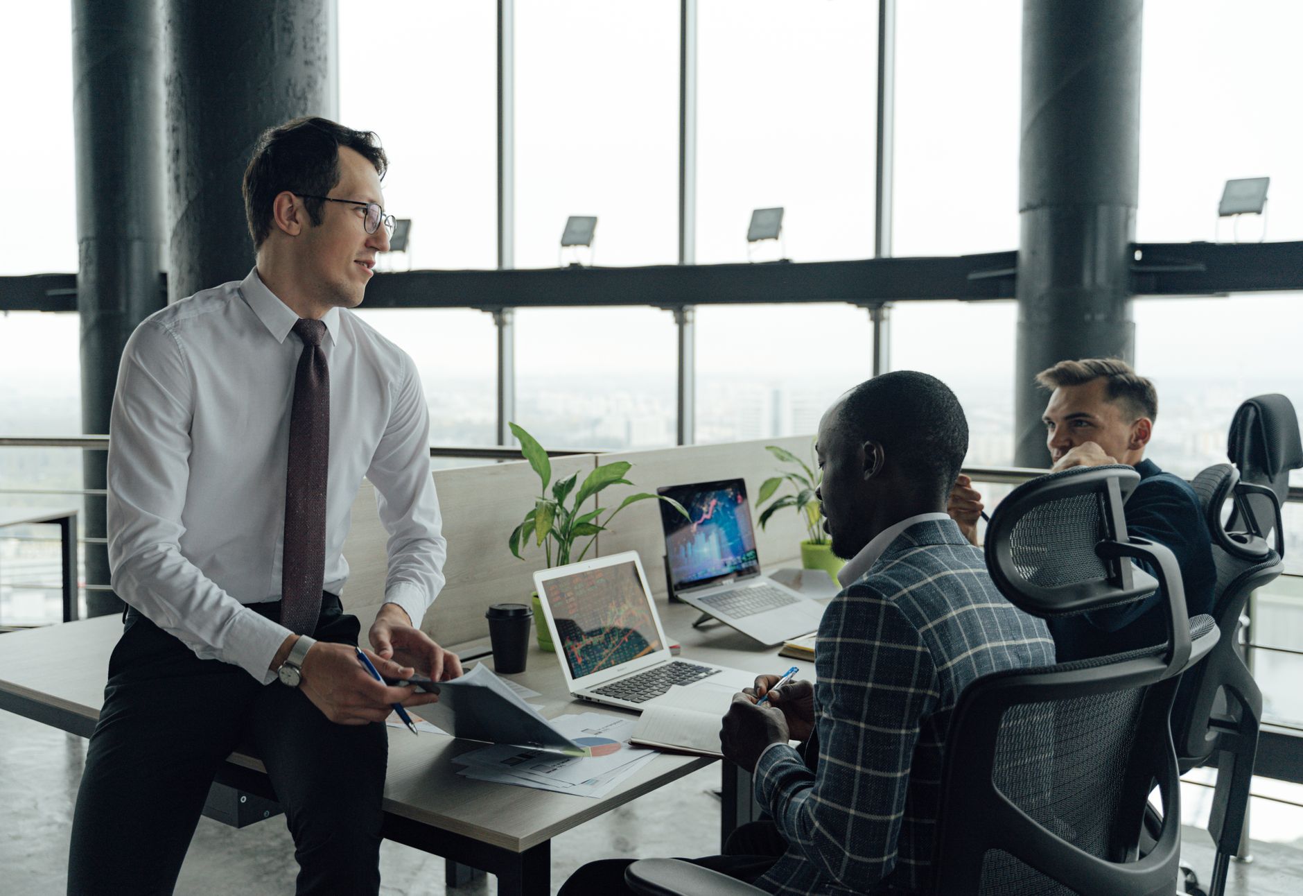 Three men engaged in a business meeting in a modern office setting with laptops and documents.