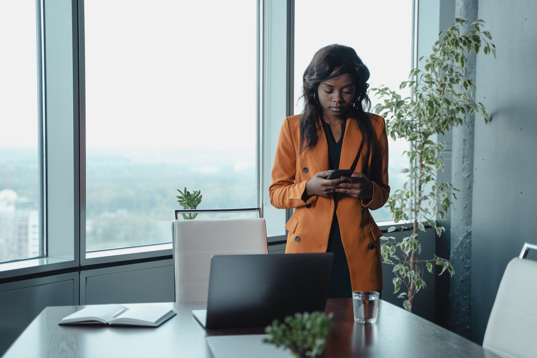 Professional woman in an orange coat checking her smartphone in an office with large windows and indoor plants.