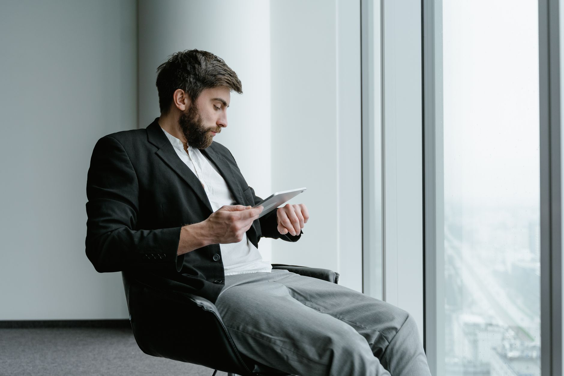 A businessman in a suit checks his watch while using a tablet in a modern office.