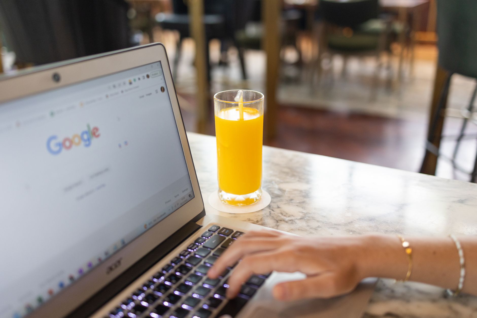 Woman typing on laptop with Google homepage open, beside a glass of orange juice on a marble table.