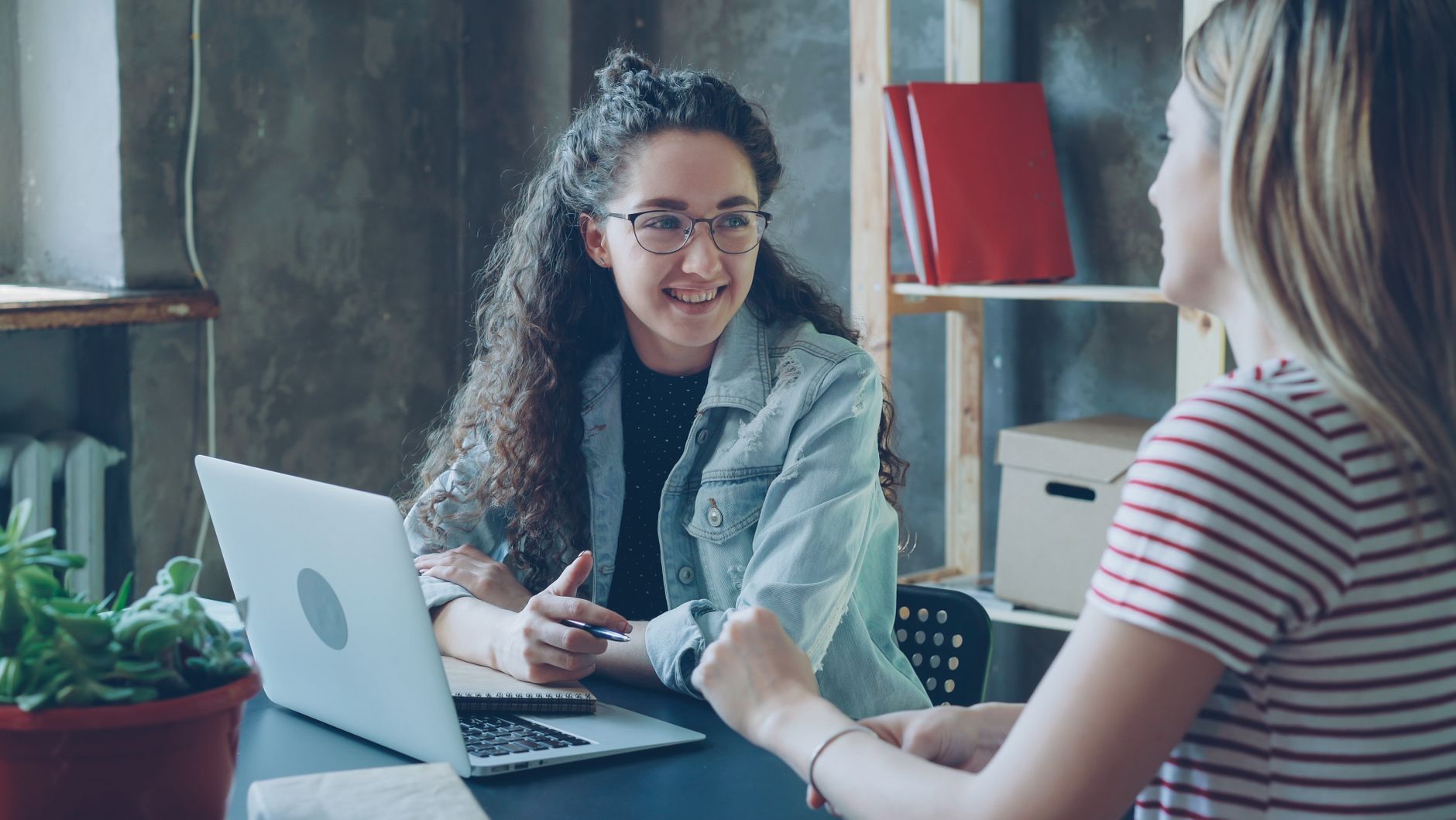 Two women engaged in a conversation at a modern office desk with a laptop.
