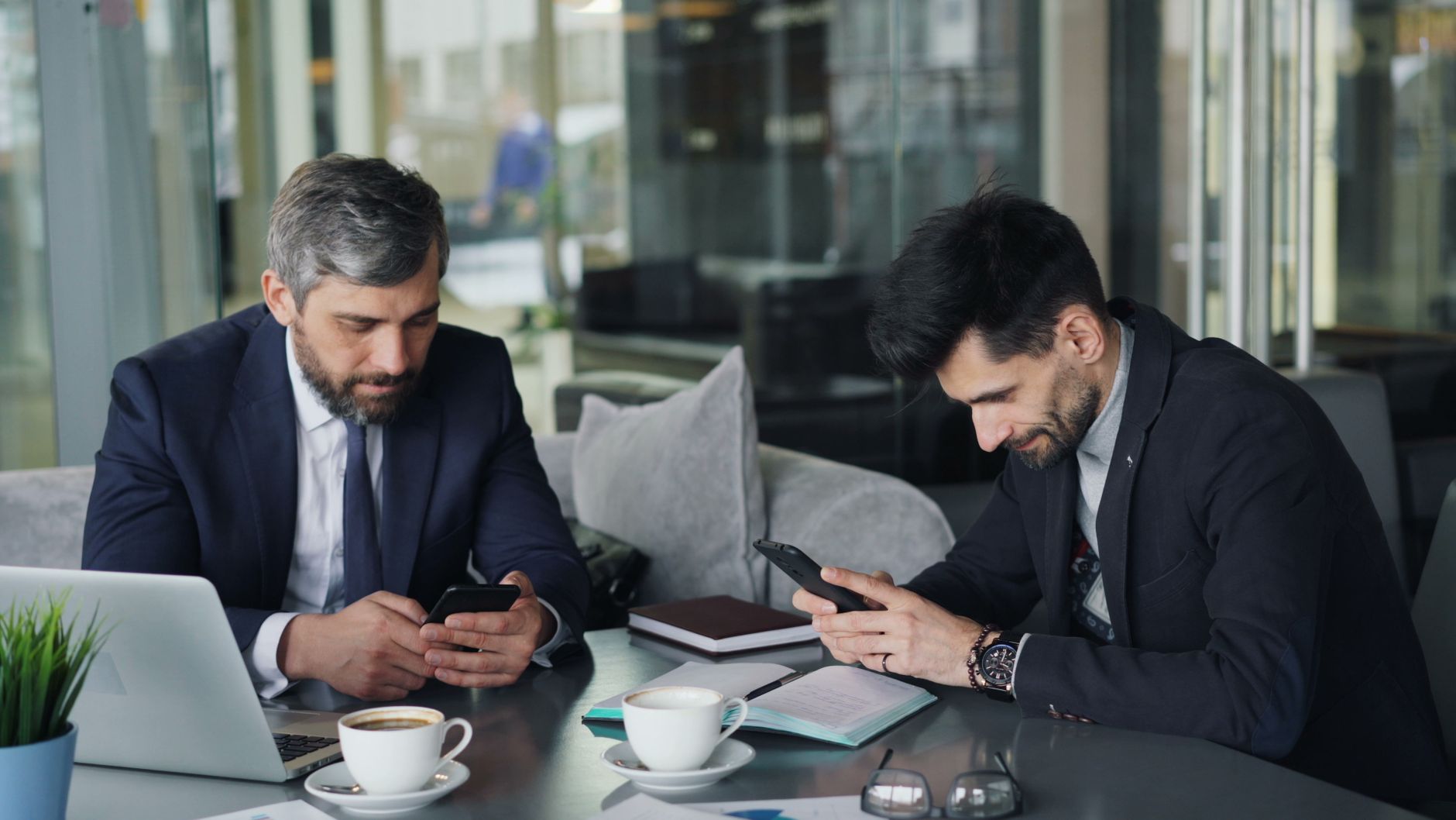 Two businessmen in suits using smartphones at a café table with laptops and coffee.