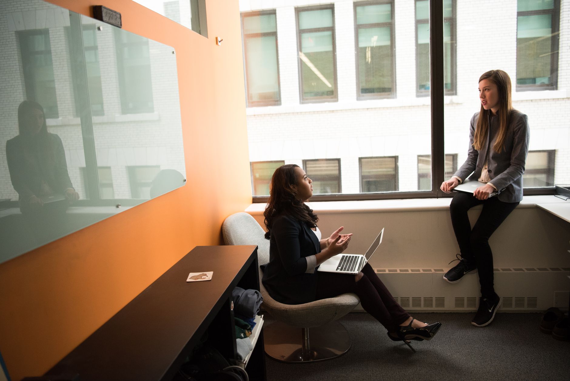 Two women engaged in a business discussion in a modern office setting with laptops and glass windows.