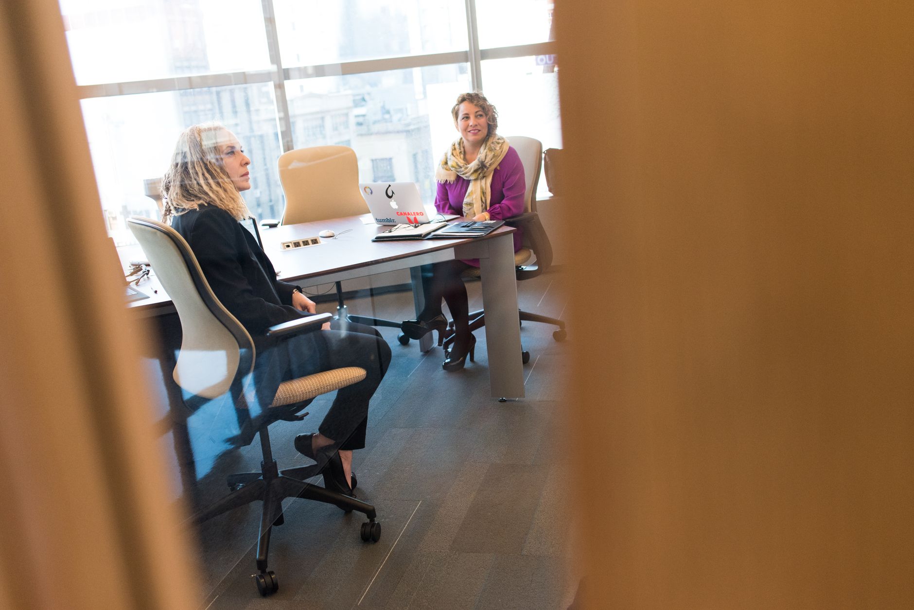 Two professional women discussing in a modern office setting with laptops and documents.