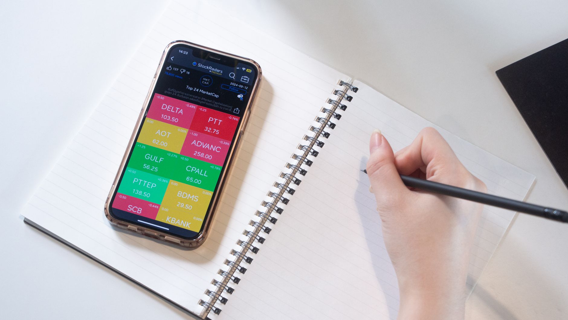 Close-up of a smartphone displaying stock data beside a hand writing in a notebook.