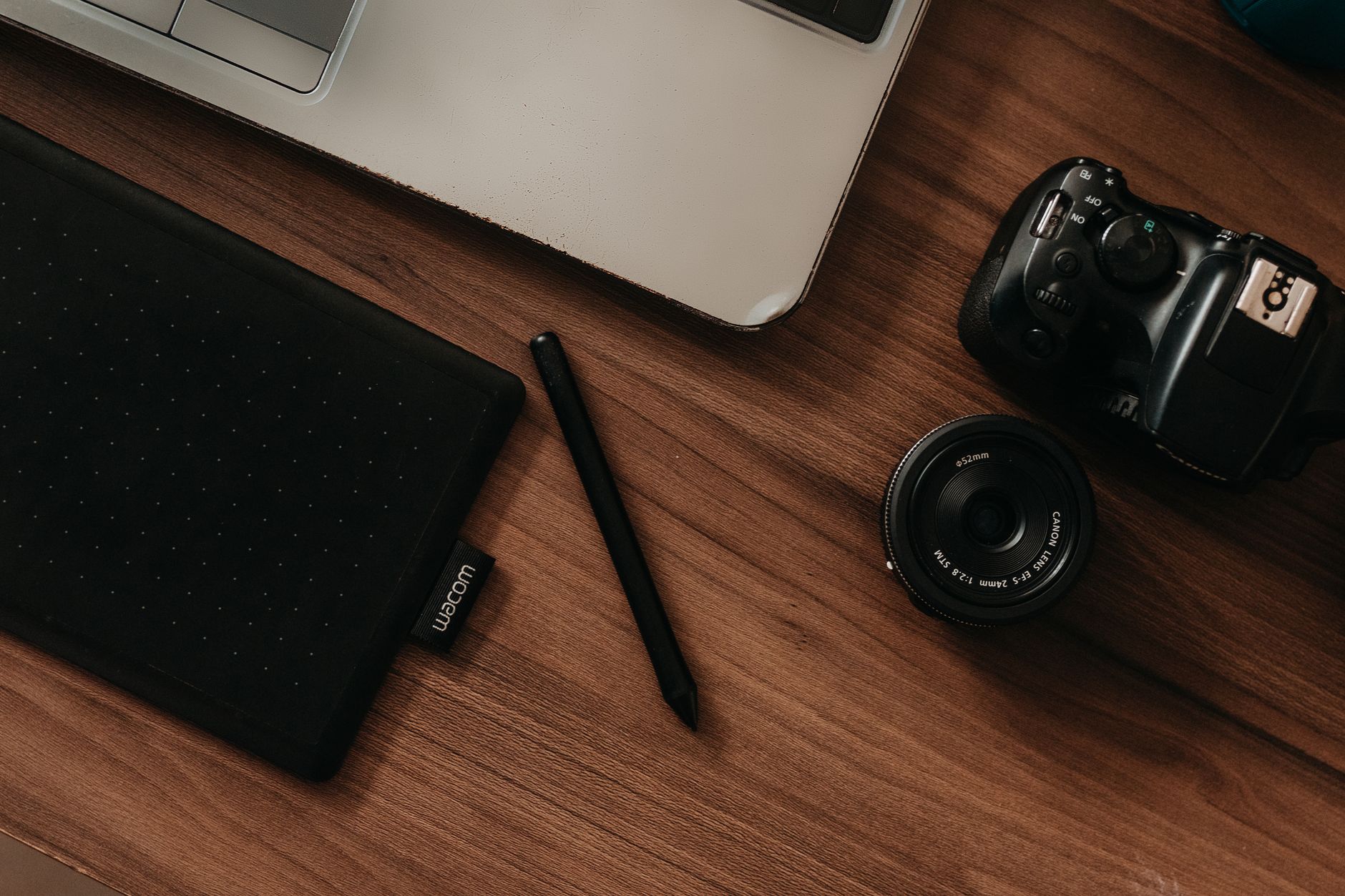 A top view of a digital workspace featuring a camera, tablet, pen, and laptop on a wooden desk.