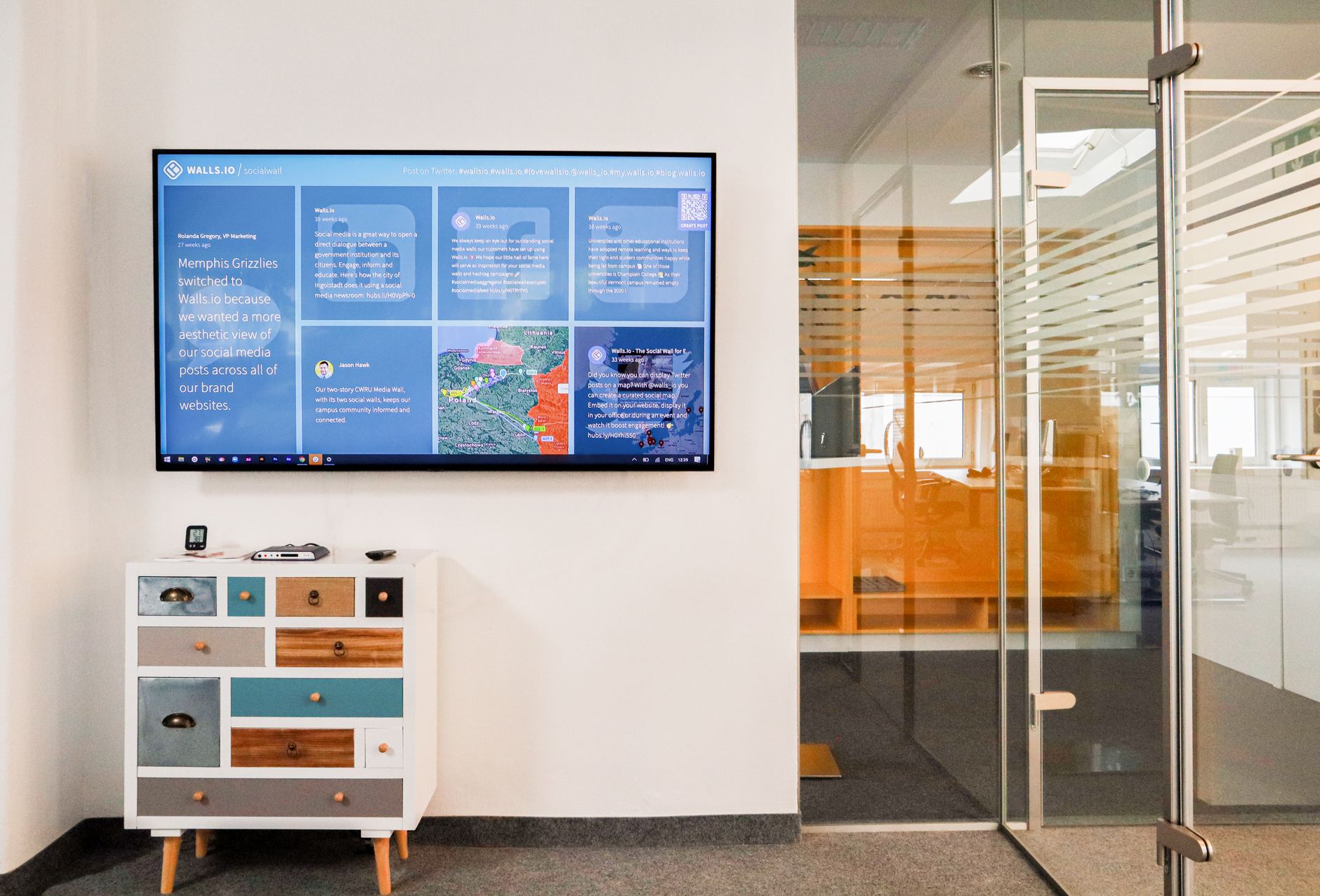 Sleek office interior featuring a wall-mounted TV screen and colorful dresser in a glass-walled room.
