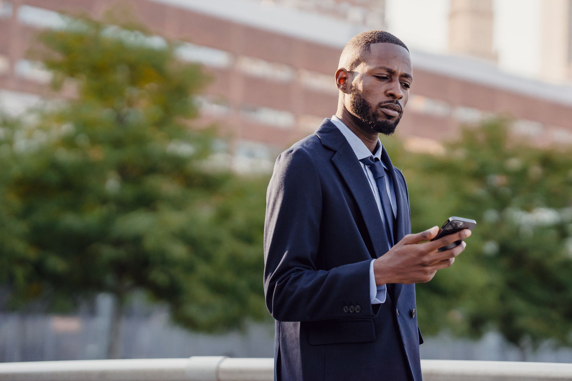 Businessman in a suit using a smartphone outdoors, surrounded by trees with a blurred building backdrop.