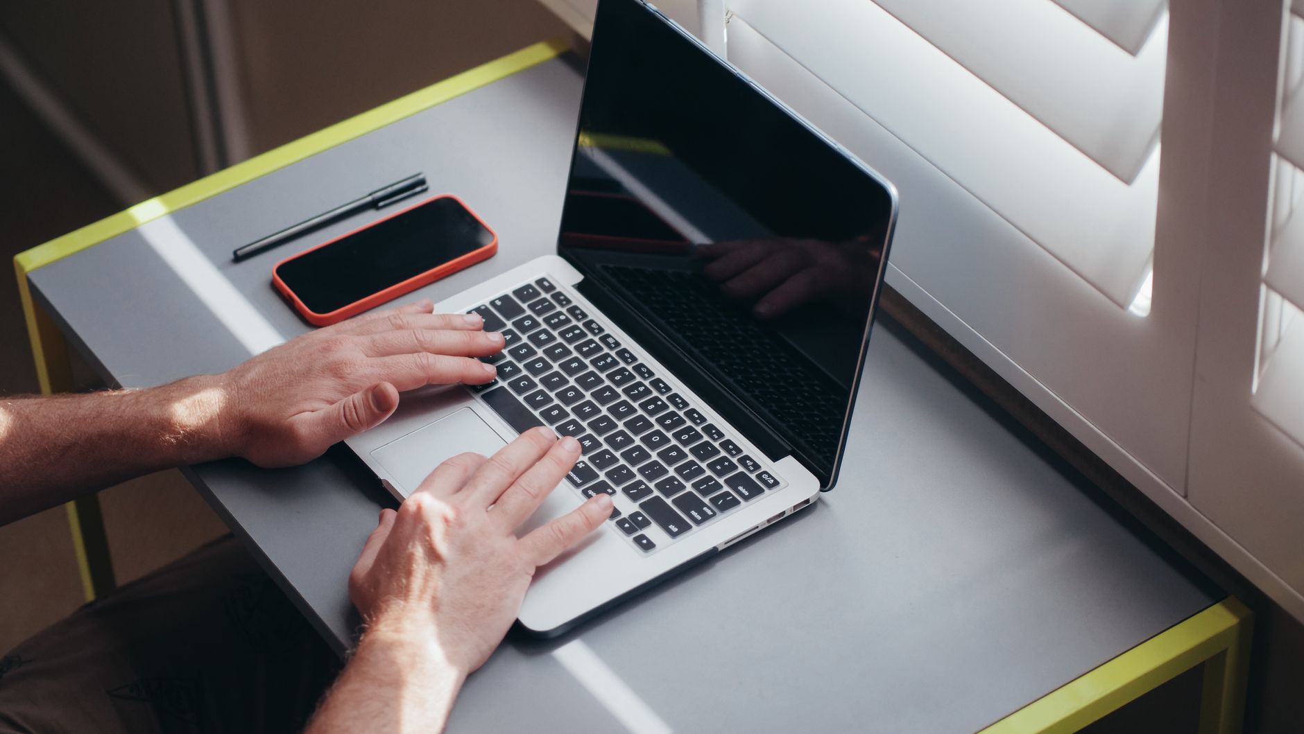 Hands typing on a laptop next to a window with a smartphone and pen on the table.