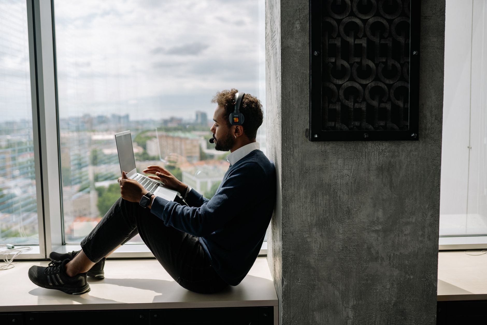 A man using a laptop while sitting by a window in an office, wearing headphones.