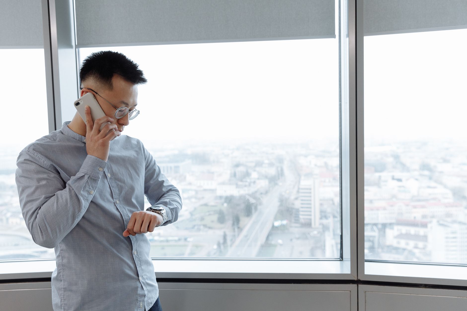 Businessman on a phone call inside a modern office, checking his watch, with a city view.
