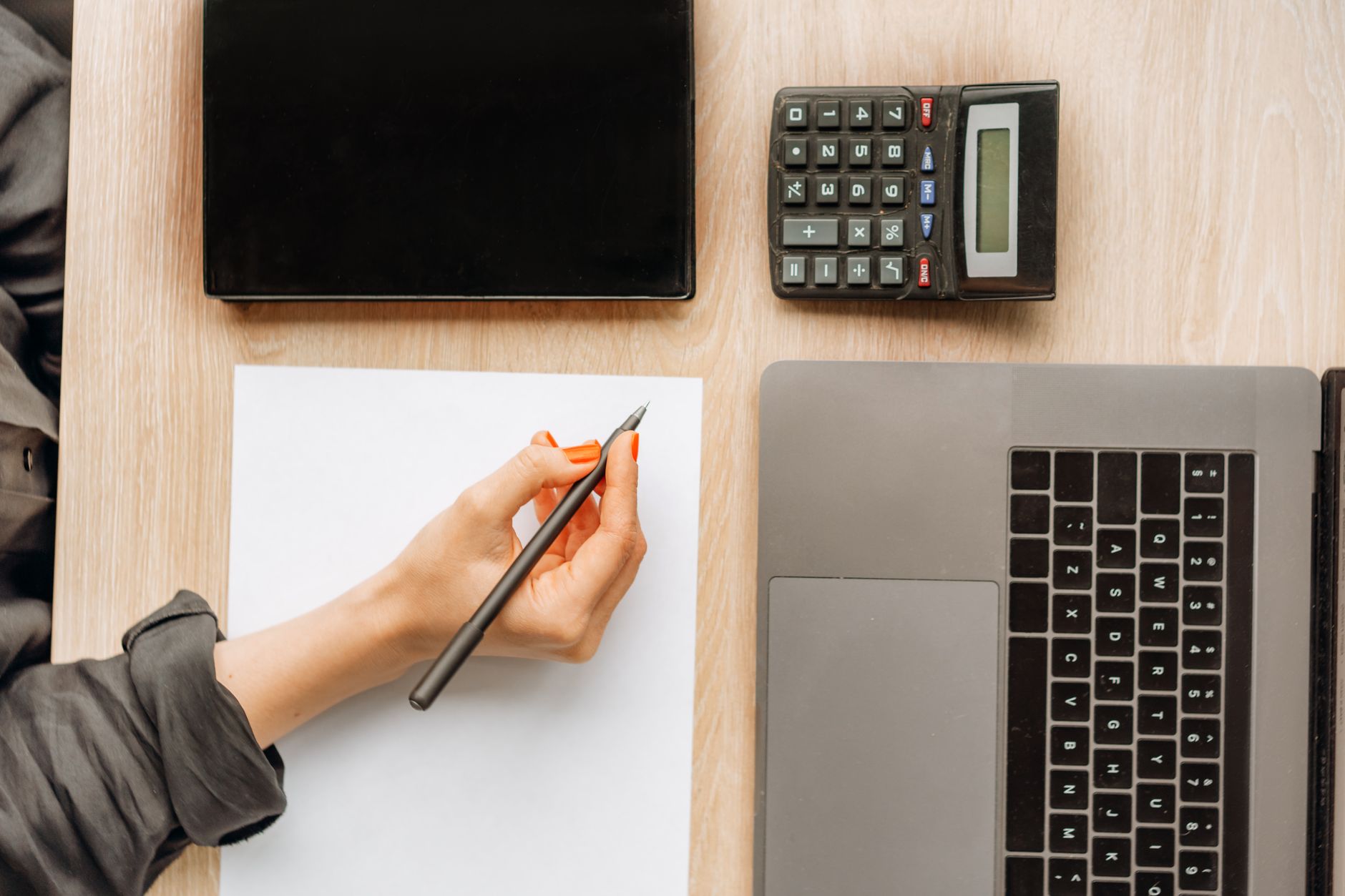 A person writing on paper surrounded by a laptop, tablet, and calculator on an office desk.