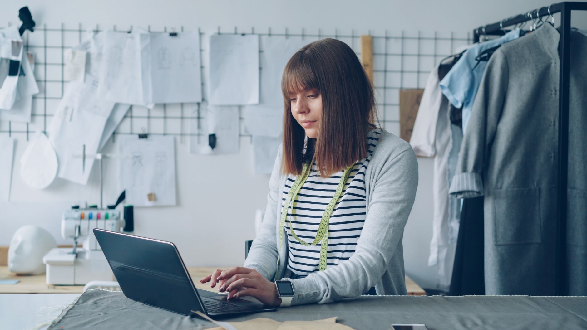 Fashion designer working on a laptop in a stylish, garment-filled workspace. Creative and focused atmosphere.