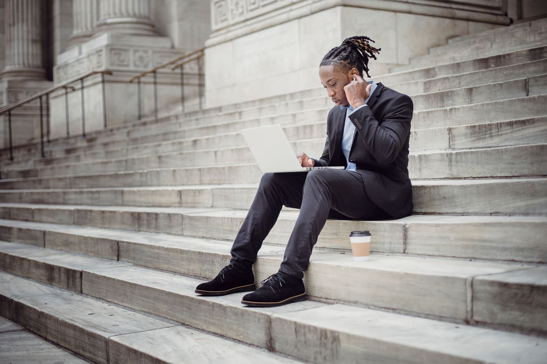 Full length focused black businessman in formal suit browsing modern netbook and using wireless earbuds while resting on stone building steps