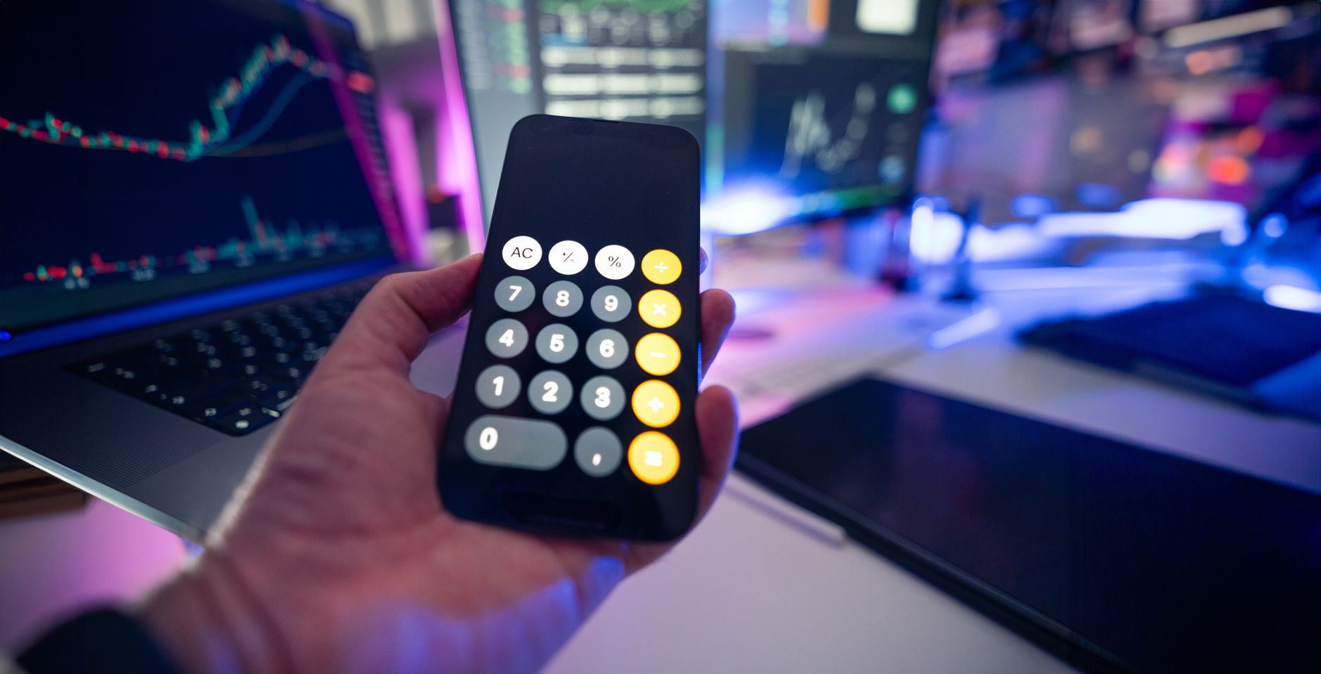 Smartphone with calculator app against financial charts on monitors, symbolizing financial planning.
