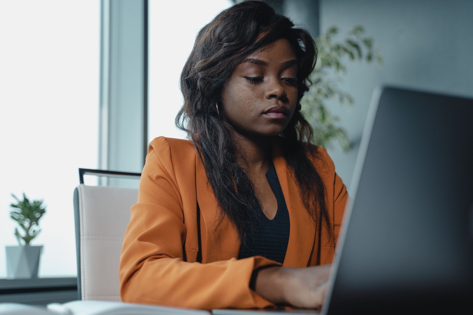 African American woman in an orange blazer working on a laptop in a modern office.