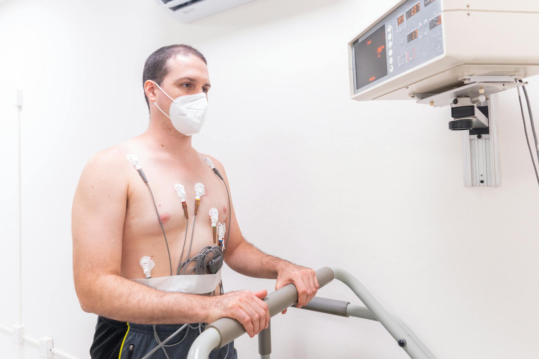 Man in medical exam wearing electrodes and face mask performing heart stress test on treadmill.