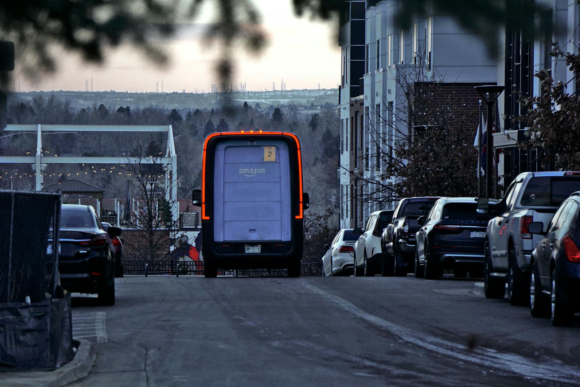 Amazon delivery van driving through an urban street at twilight with cars parked along the side.