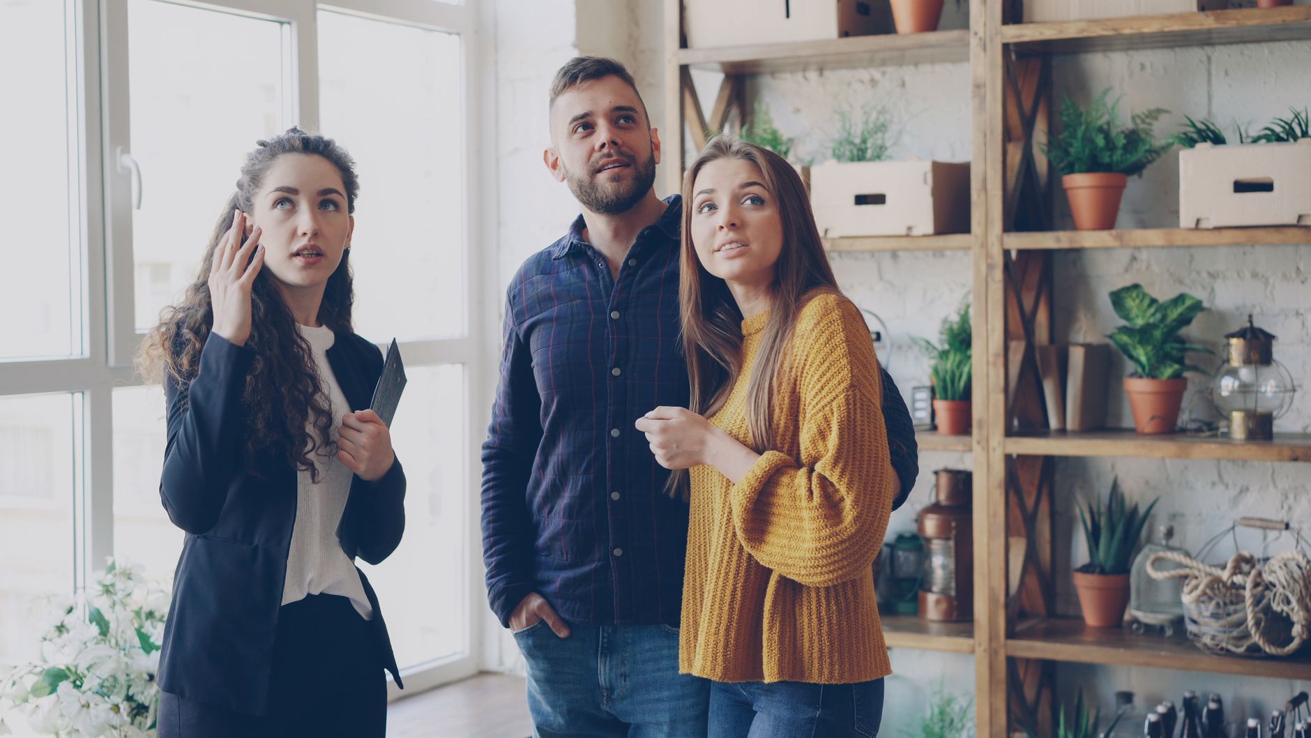 Three young adults in a stylish indoor setting with plants and shelves, engaged in conversation.