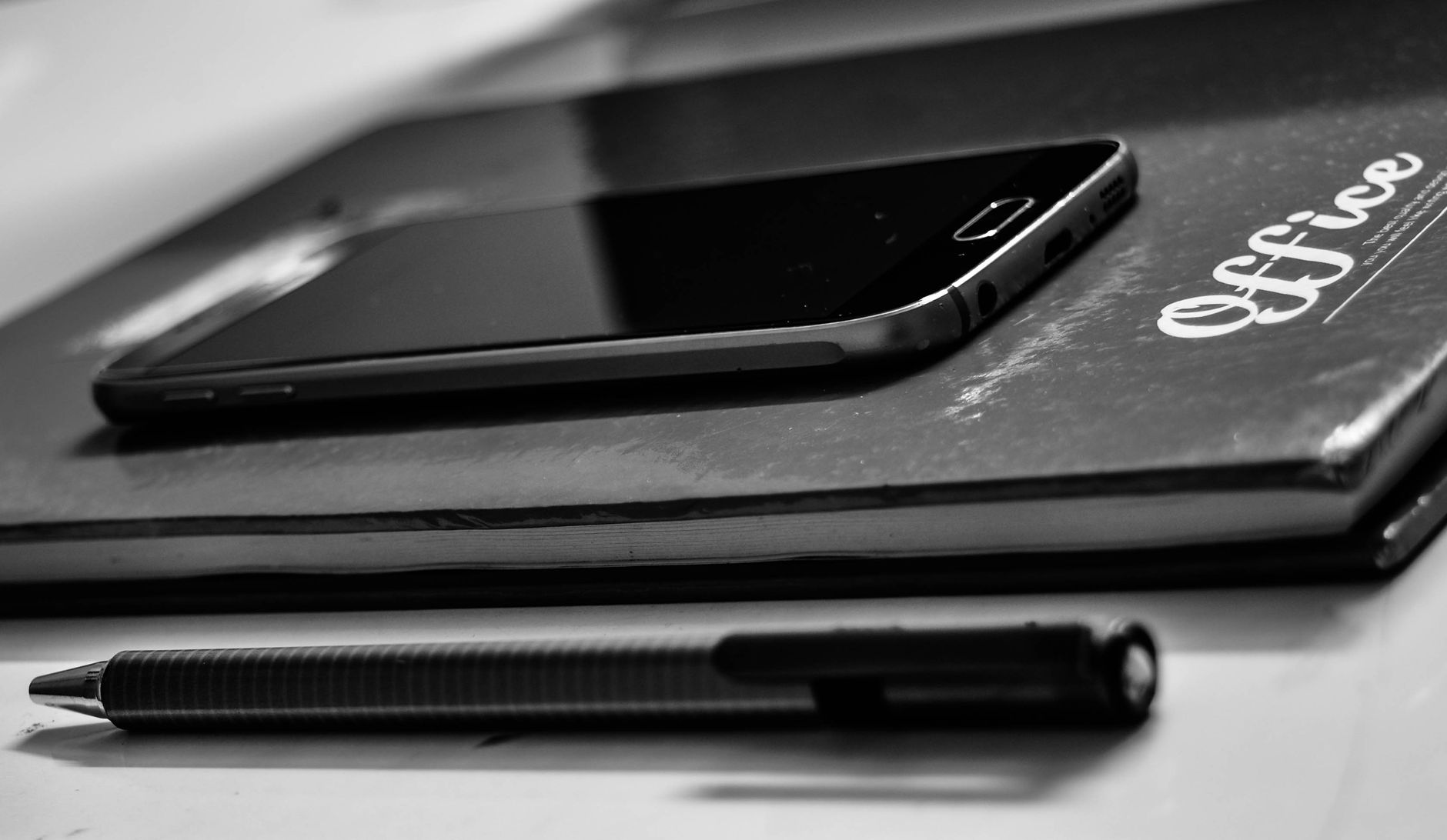 Monochrome close-up of a smartphone, notebook, and pen on an office desk.