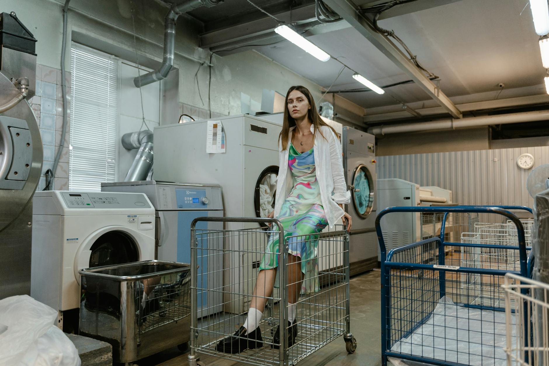 A woman in a colorful dress sits on a laundry cart in a busy industrial laundry facility.