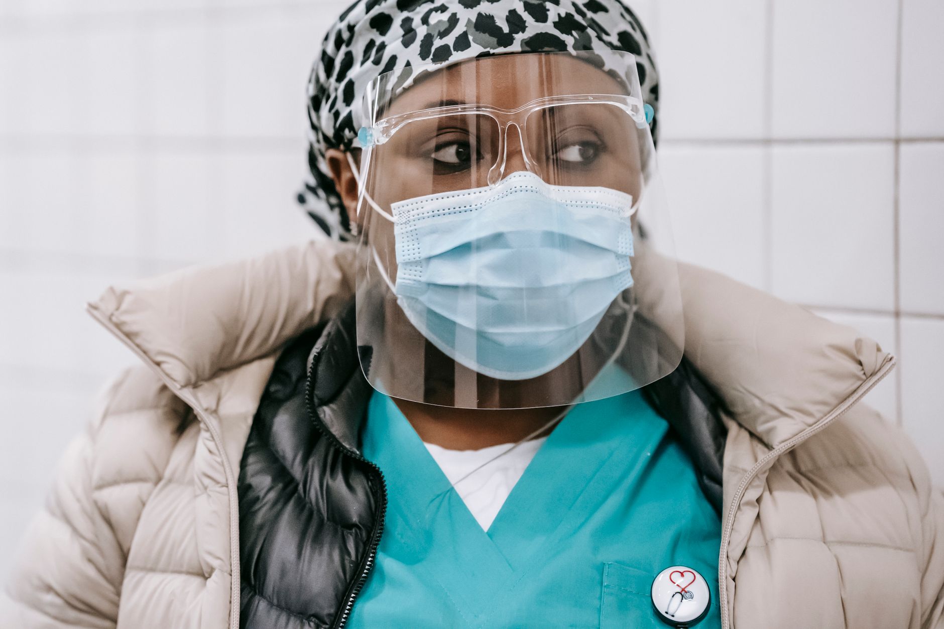 Crop contemplative African American female doctor in uniform and warm clothes wearing face mask and shield standing against white tile wall and looking away