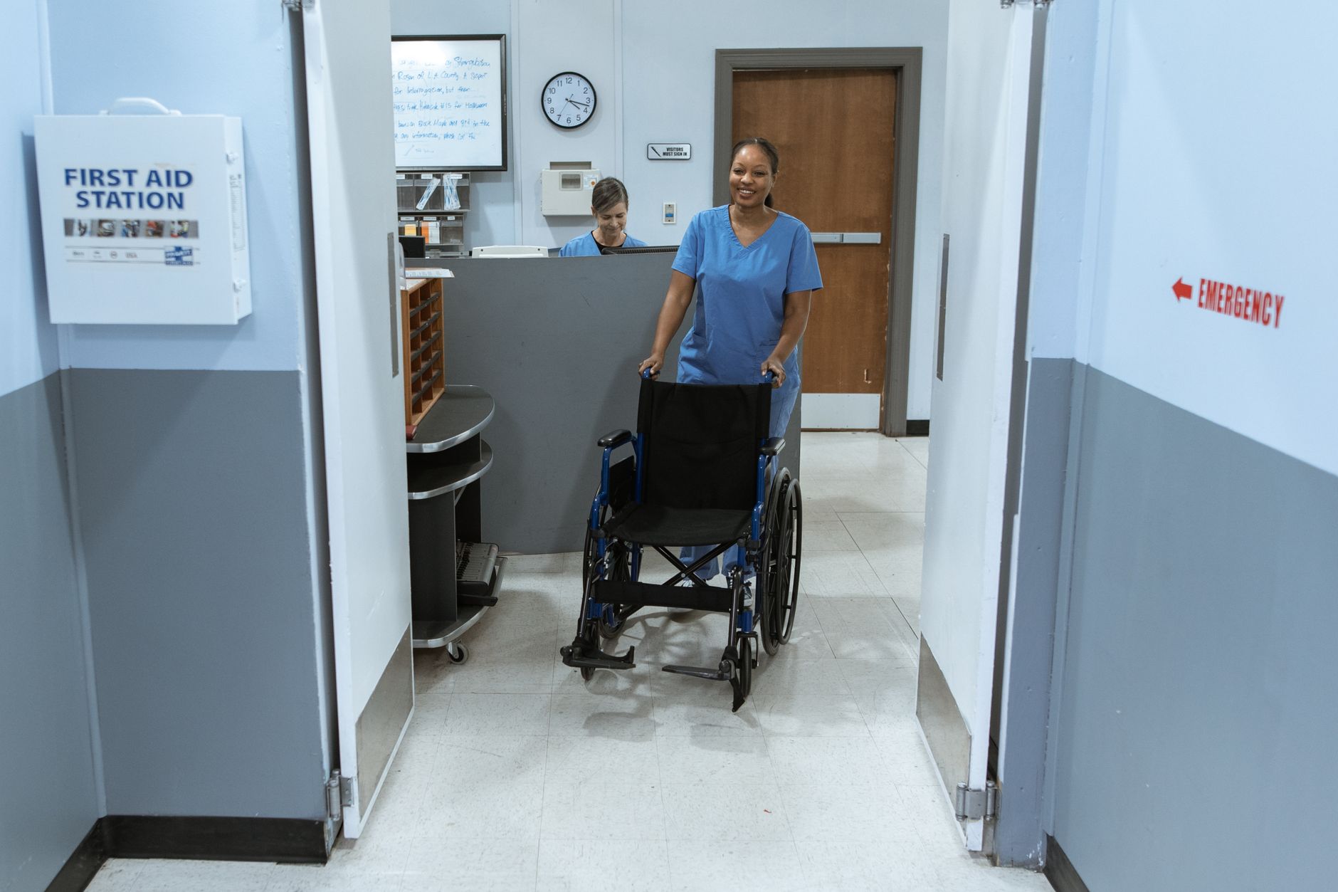 A nurse in scrubs pushes an empty wheelchair through a hospital hallway.