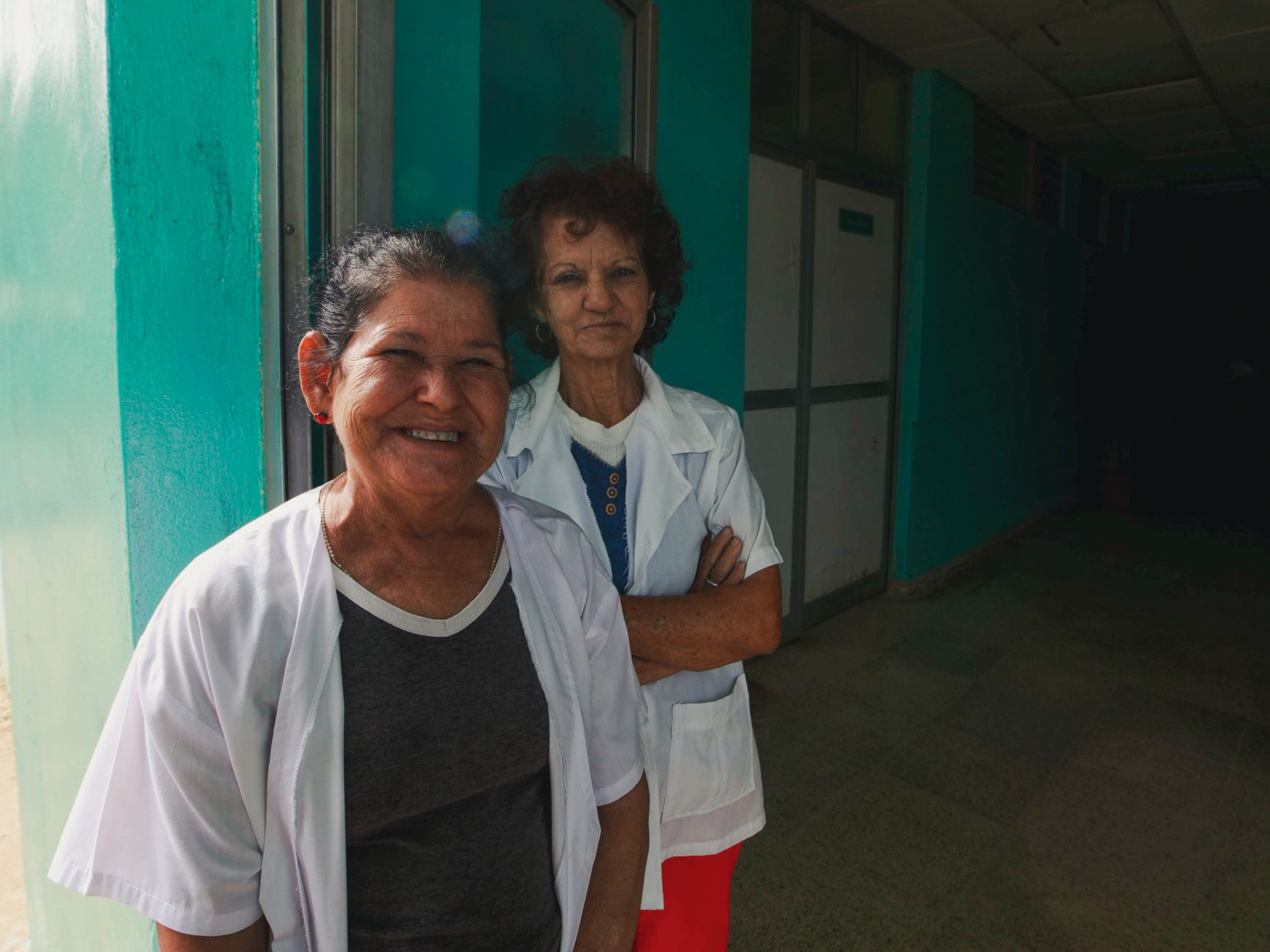 Two women healthcare workers smiling in a hospital corridor, wearing uniforms.