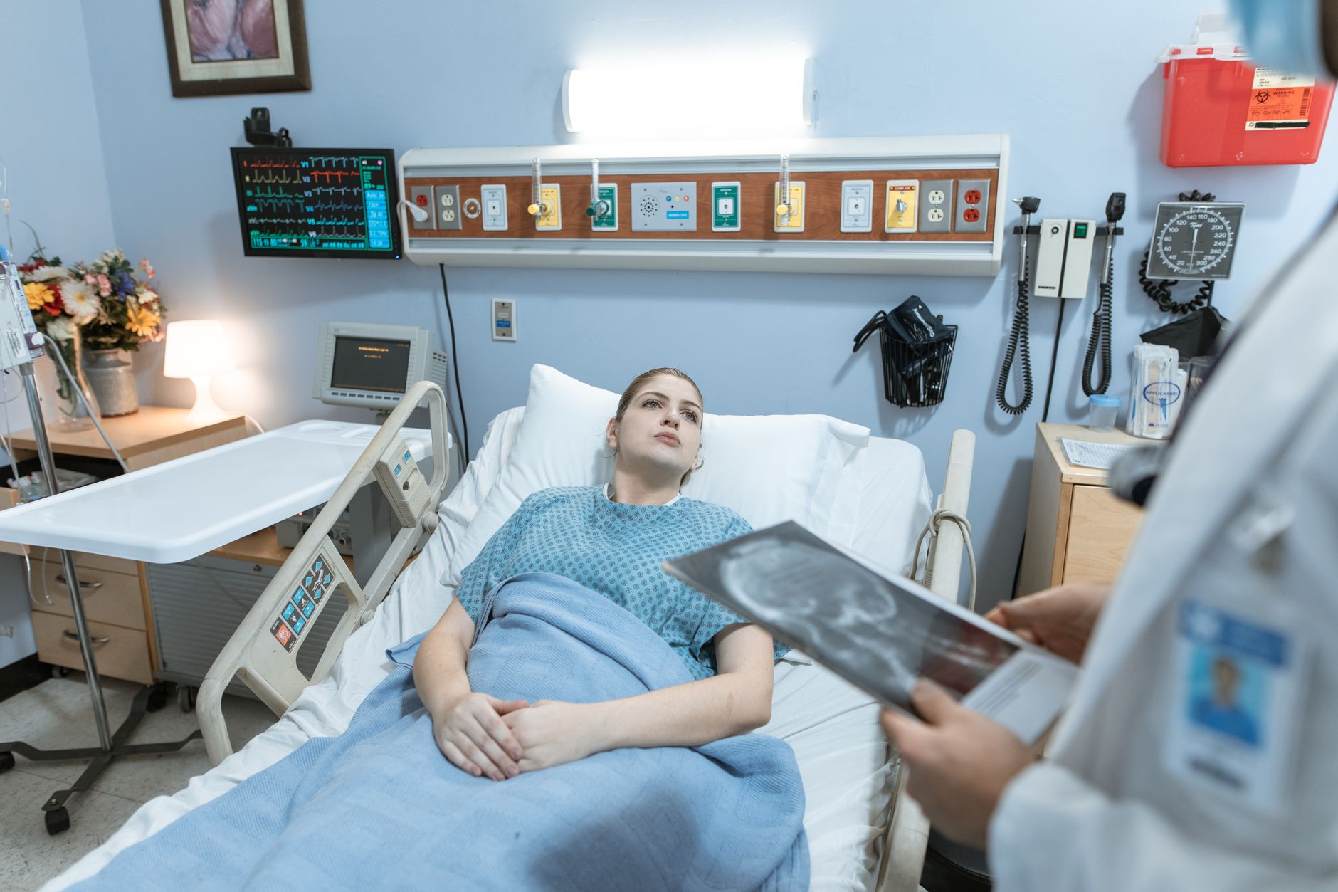 A female patient in a hospital gown receives medical attention from a healthcare professional holding an x-ray in a hospital ward.