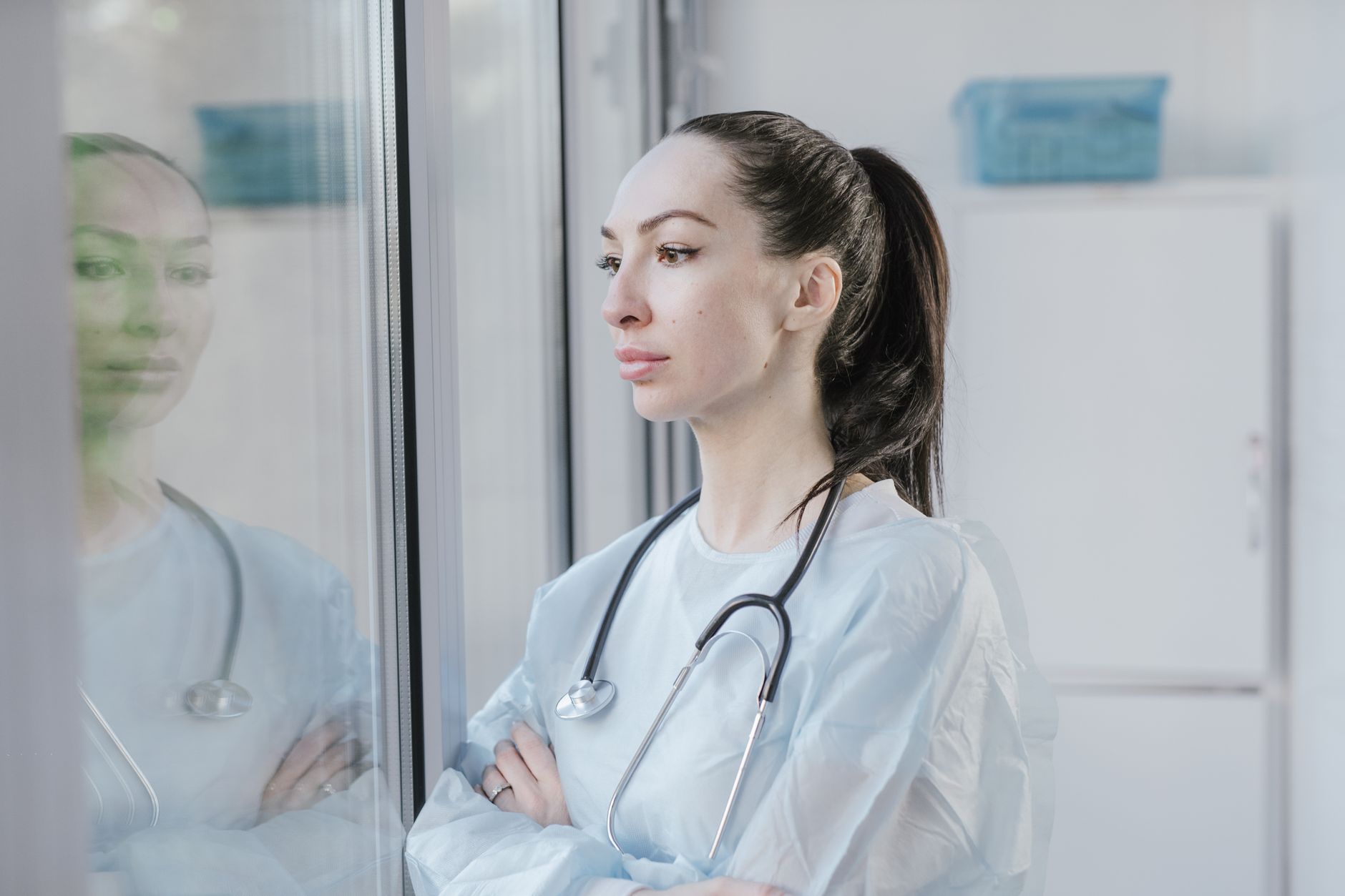 Female doctor in a lab coat with stethoscope, pondering while looking out a window.
