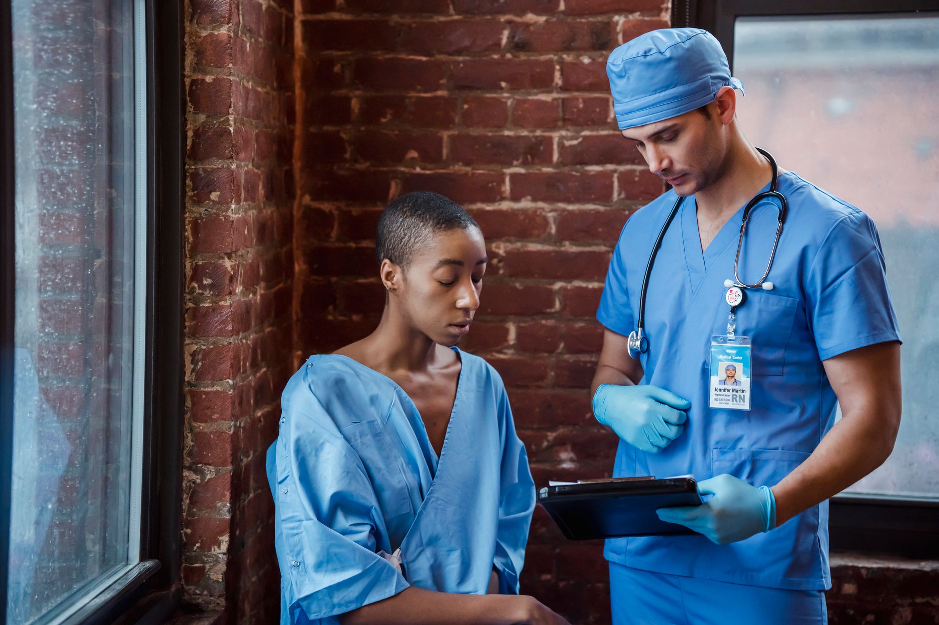 Pensive man nurse in blue uniform standing and demonstrating diagnosis to serious African American female patient in hallway of hospital in daytime