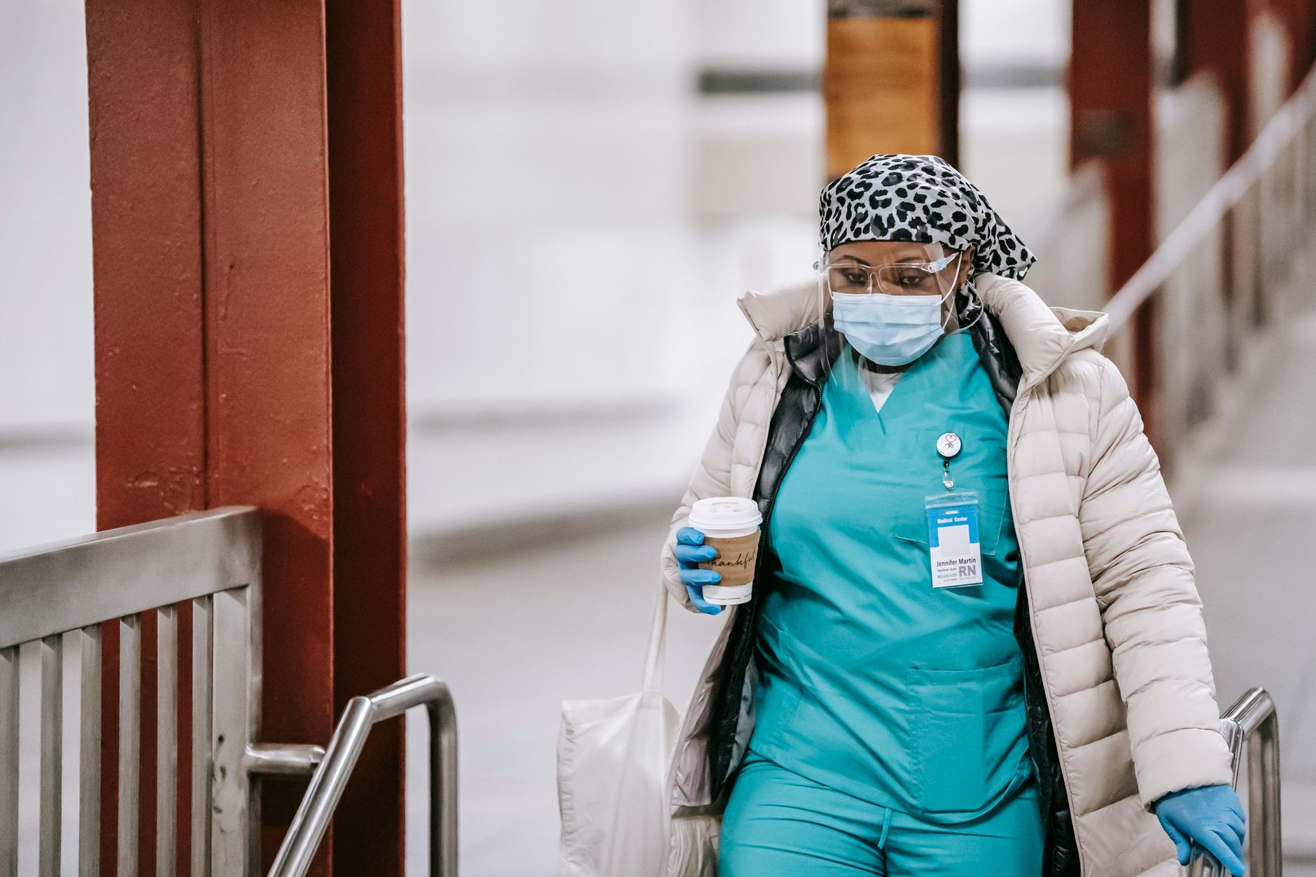 Serious female medical specialist wearing uniform protective mask and gloves walking on street with paper cup with hot drink