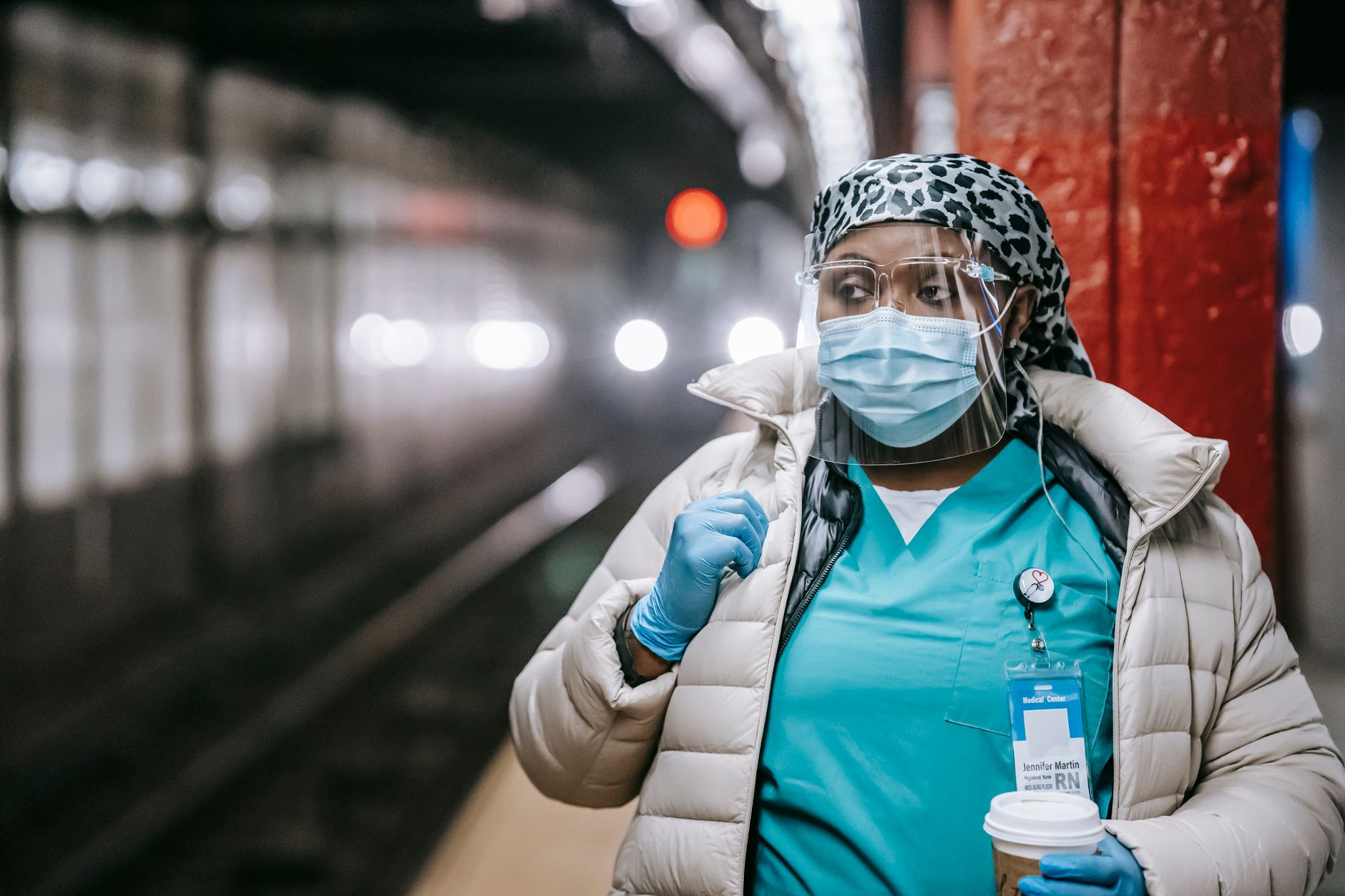 Serious plump African American nurse in uniform and outerwear wearing face mask and latex gloves standing with takeaway coffee standing on metro station platform and looking away thoughtfully