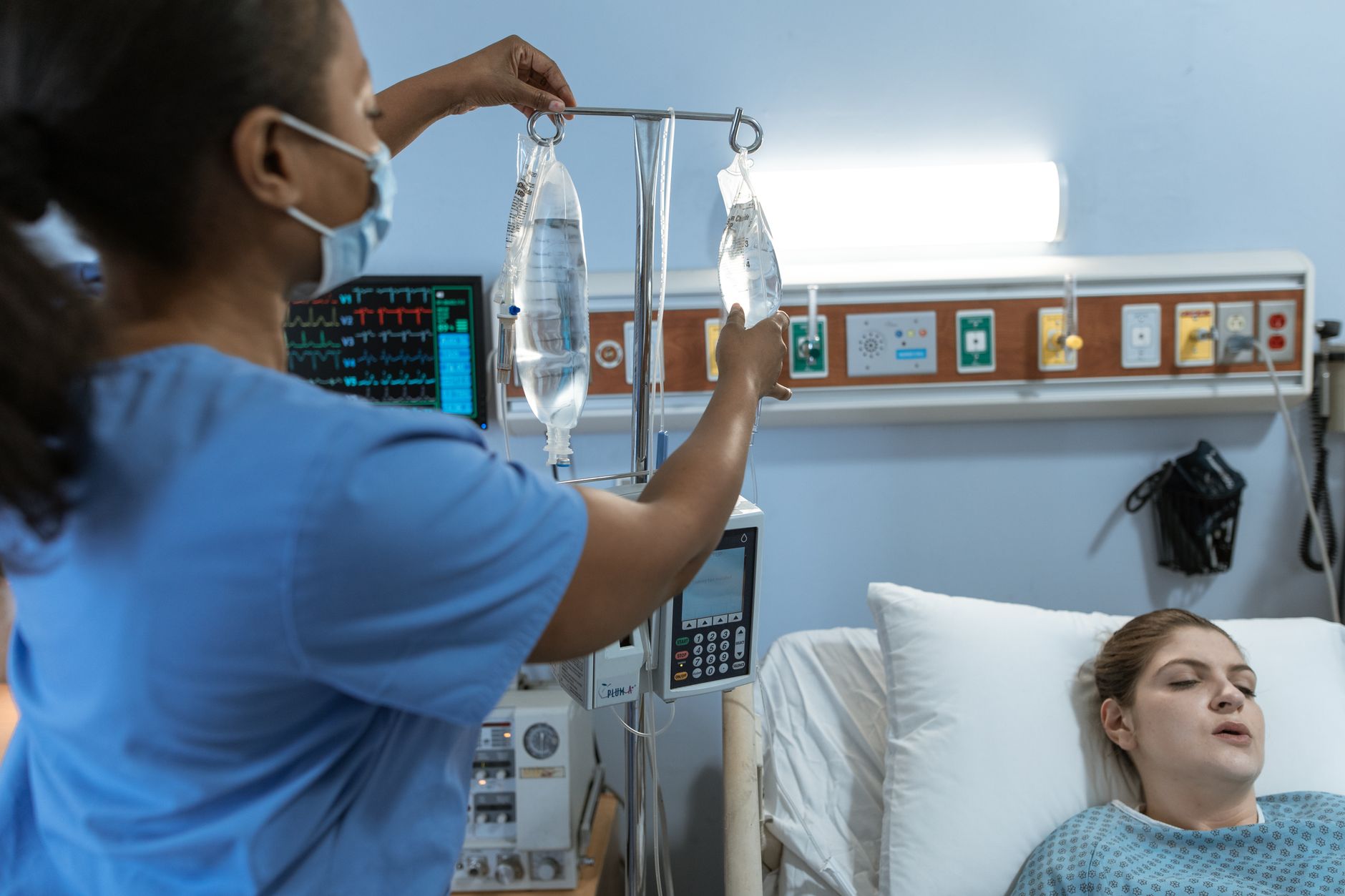 A nurse in a hospital setting adjusts an IV drip for a patient lying in bed, showcasing medical care.