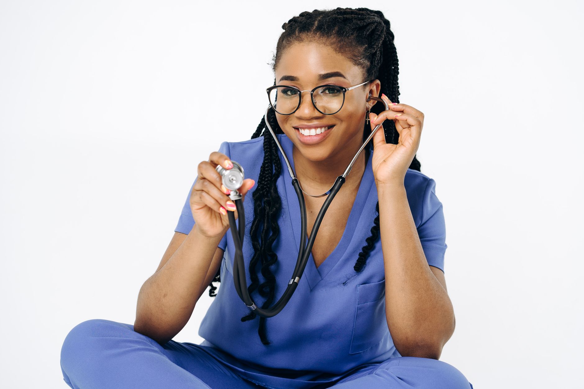 Confident female doctor in blue scrubs smiling and holding a stethoscope against a white background.