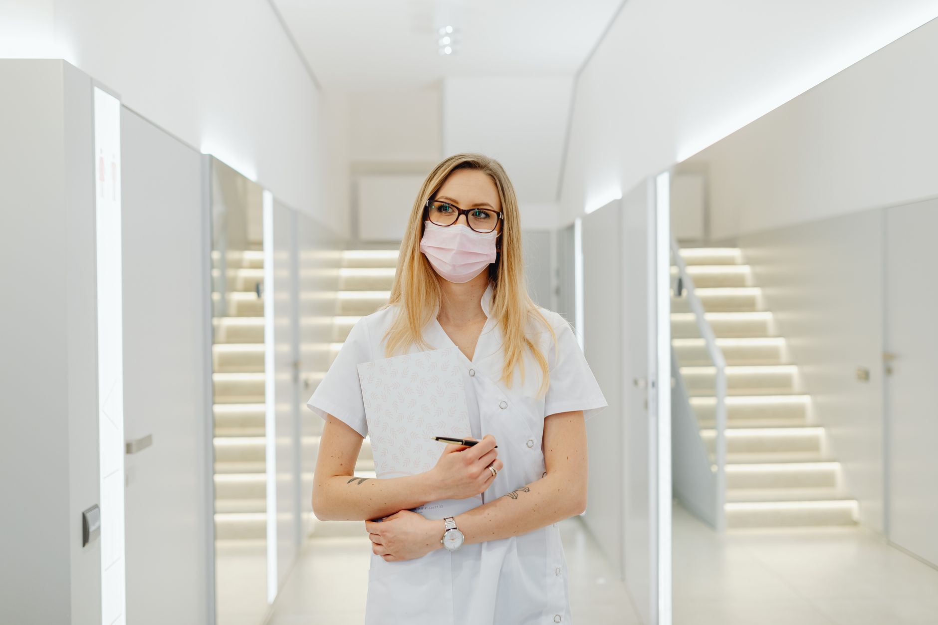 A woman healthcare worker wearing a mask and holding documents in a modern clinic hallway.