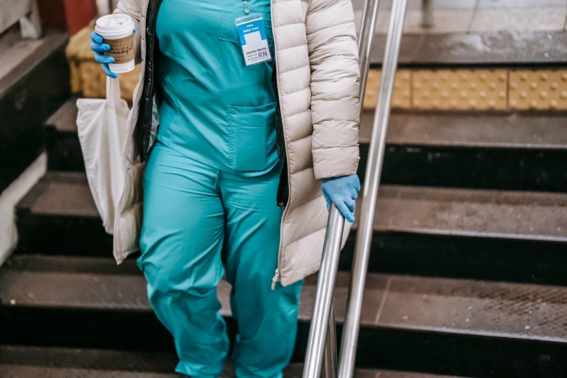 Crop faceless nurse in outerwear and latex gloves going with coffee to go downstairs in pedestrian passage during coronavirus period