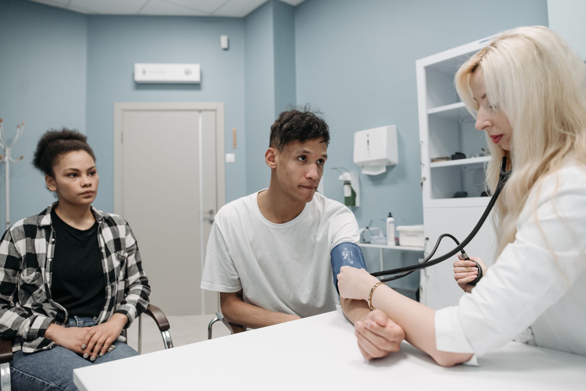 A doctor checks a patient's blood pressure while a companion observes in a clinical setting.