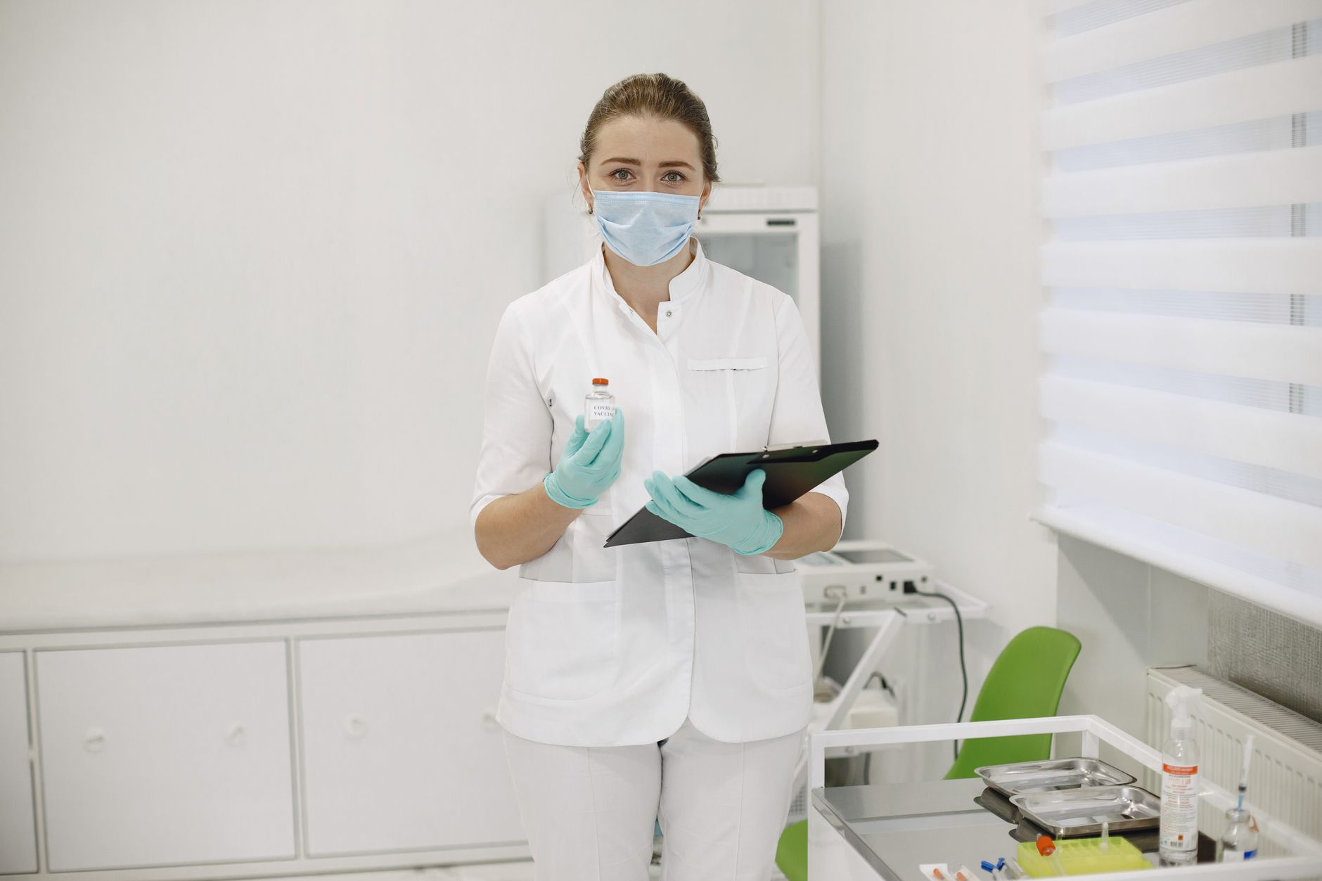 Medical professional in protective gear holding vaccine vial and clipboard in a clinical setting.