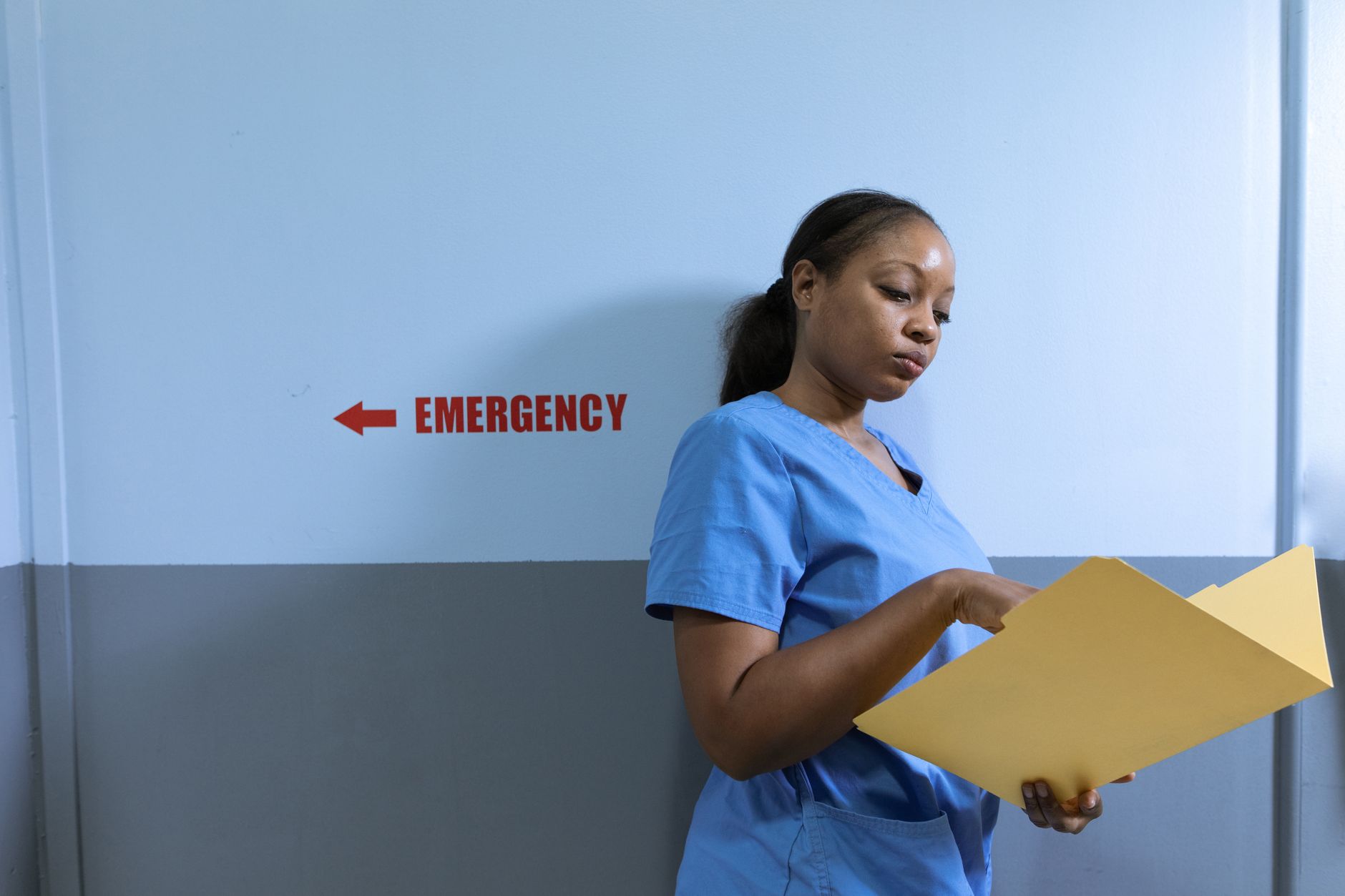 A nurse in blue scrubs reads a folder near an emergency room sign in a hospital.