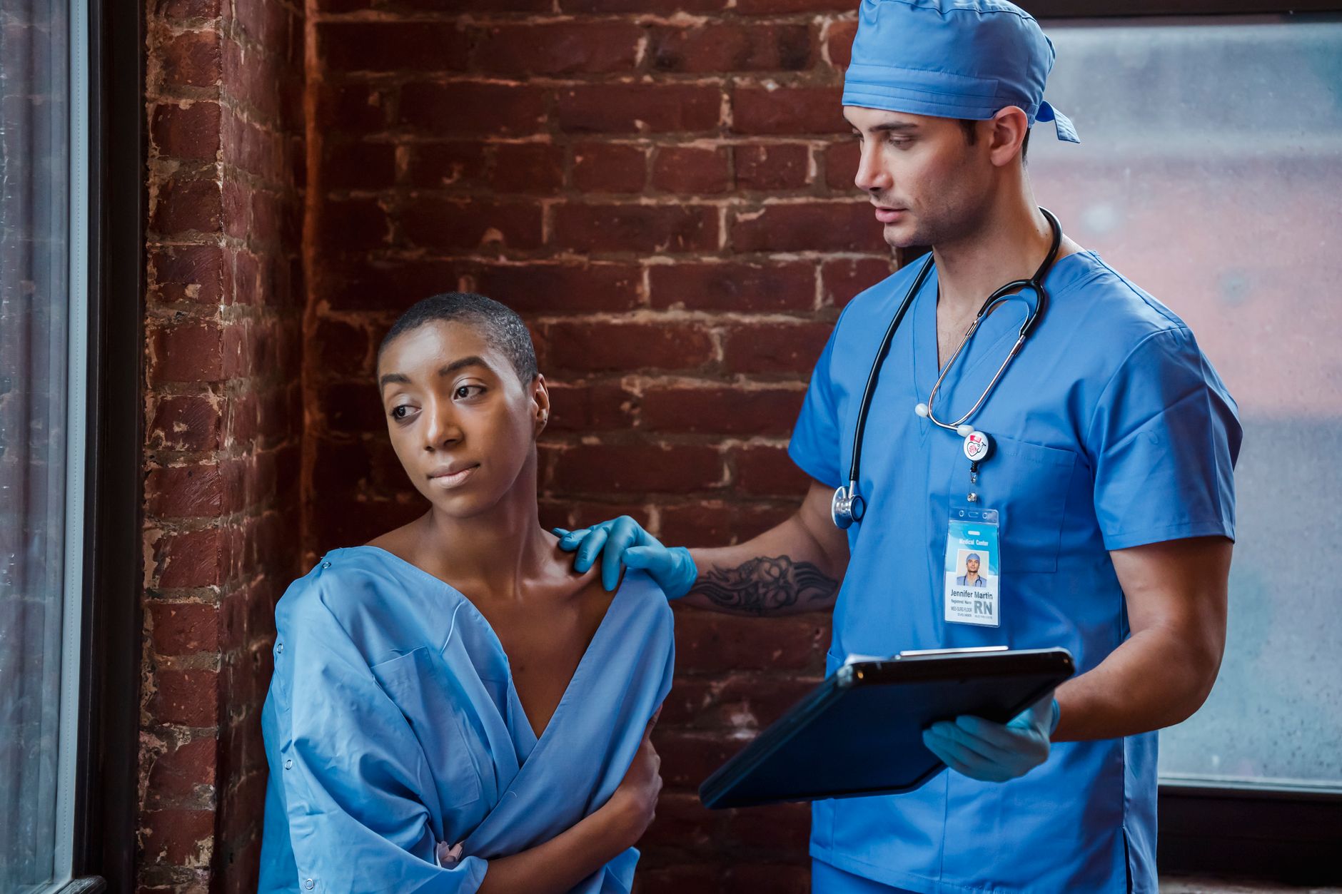 Man doctor in blue uniform and sterile gloves with stethoscope demonstrating diagnosis to thoughtful African American female patient in corridor of clinic in daytime