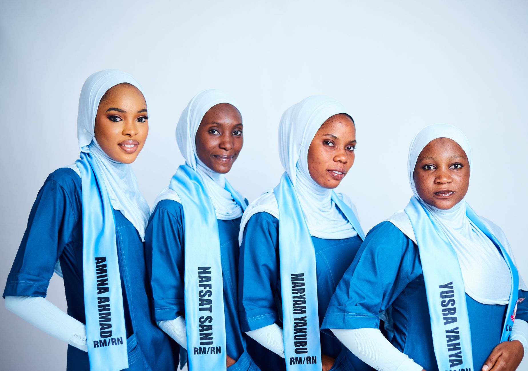 Four nurses in hijabs and blue uniforms, smiling against a plain background.