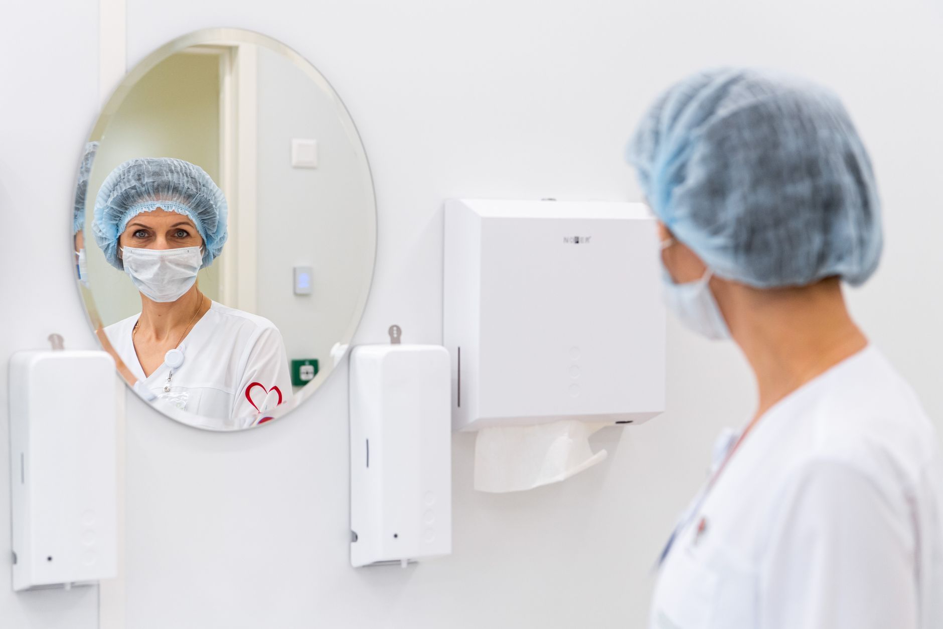 A female doctor wearing surgical scrubs and a mask looks at her reflection in a hospital mirror.