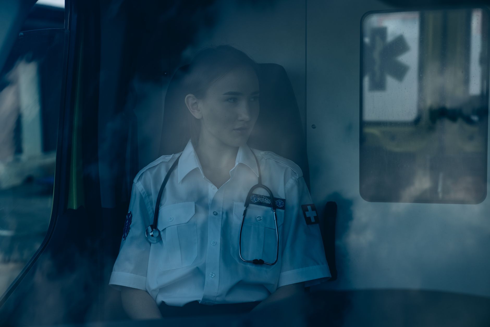 A thoughtful paramedic sits inside an ambulance, wearing a uniform and stethoscope.