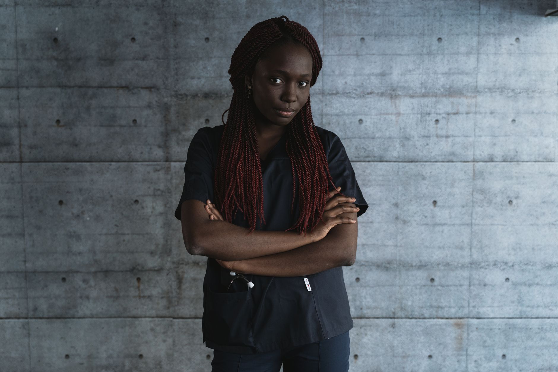 Portrait of a confident female medical professional with braided hair, posing with crossed arms against a concrete wall.