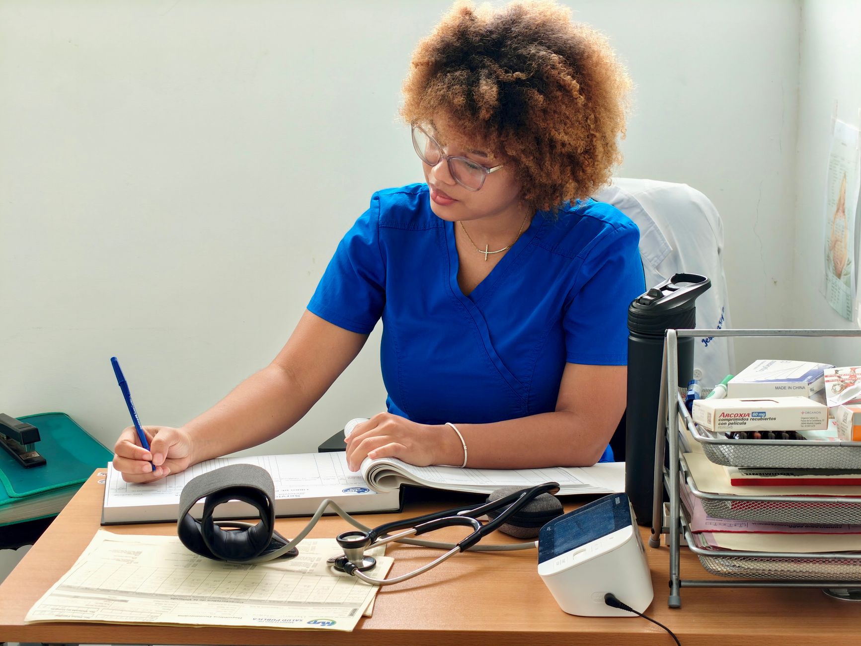 A nurse in blue scrubs writing patient information at a desk with medical equipment.